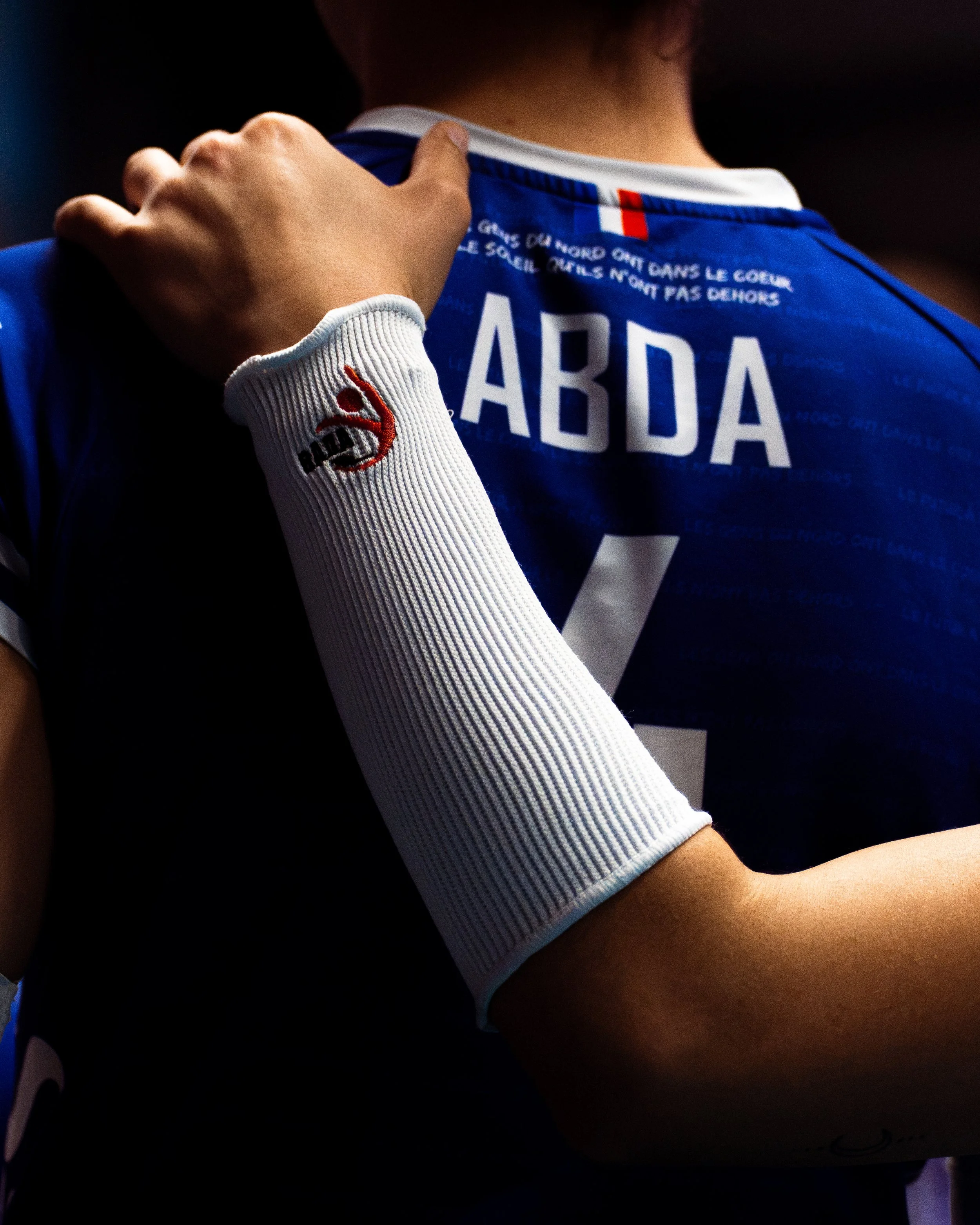 Un joueur de football portant un maillot bleu avec le nom « ARDA » visible en arrière-plan, et une manche blanche avec un logo rouge et noir, en train d'être assisté ou ajusté par une autre personne.