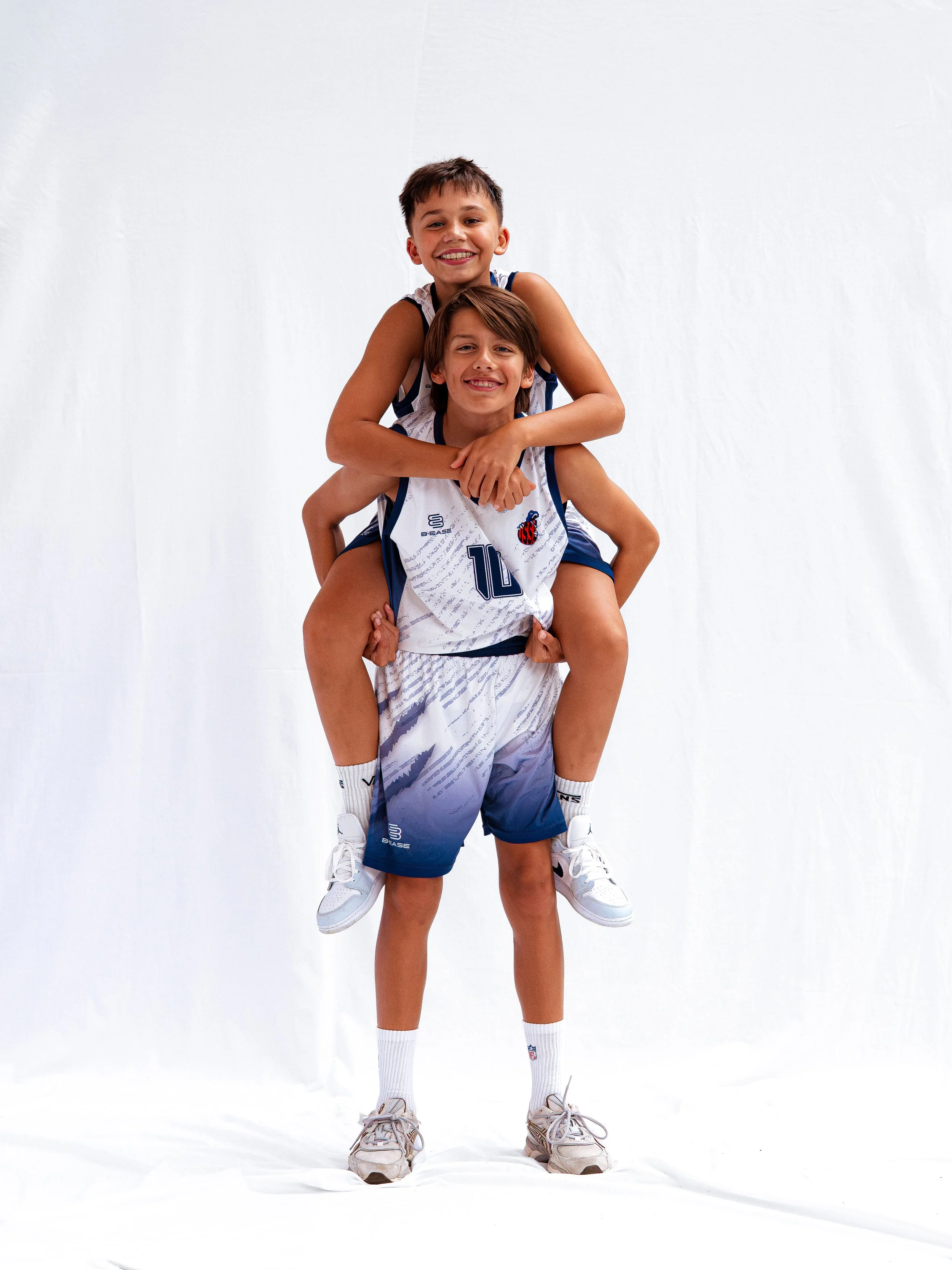 Deux jeunes garçons portant des maillots de basketball, l'un sur les épaules de l'autre, souriant devant un fond blanc.