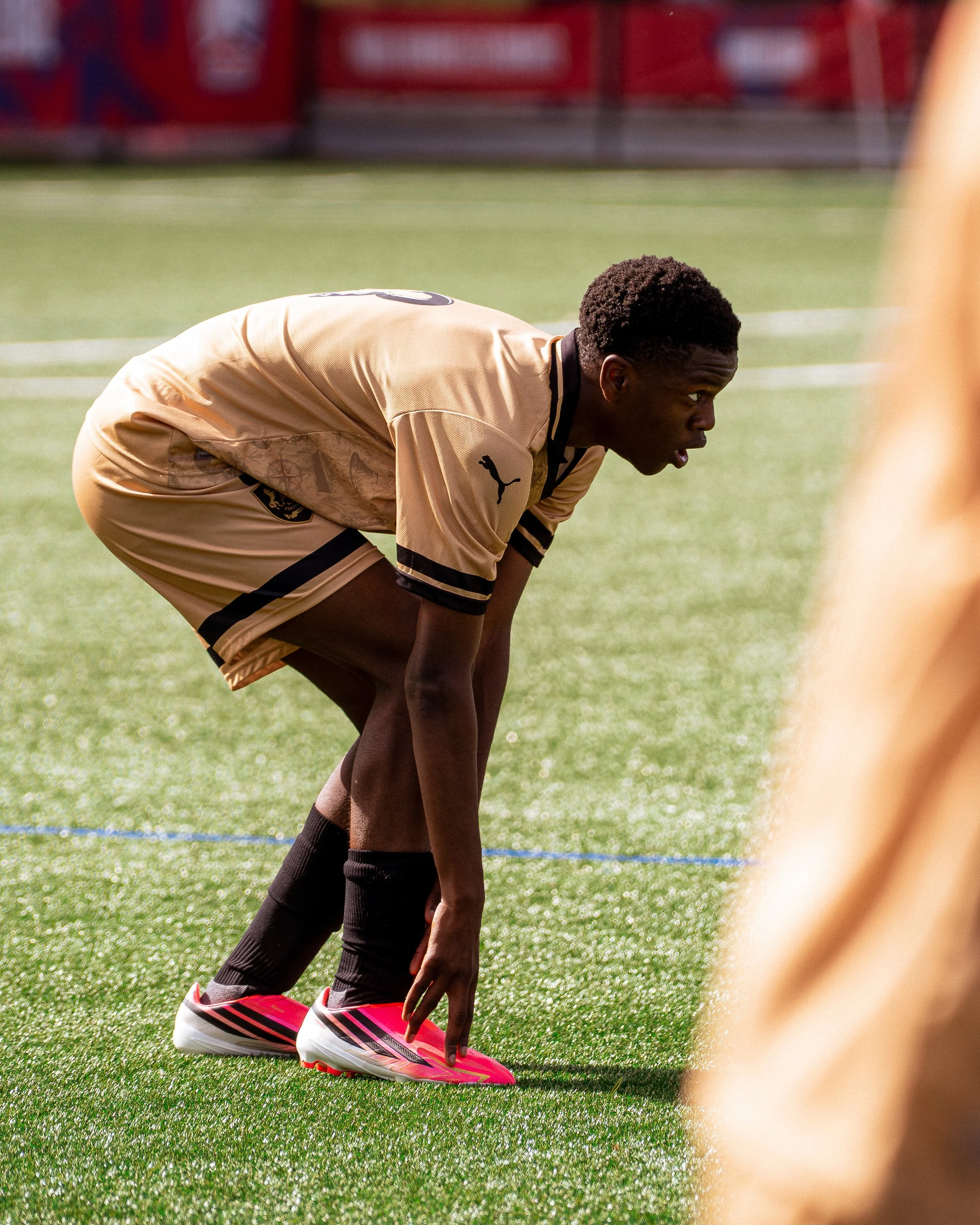 Un joueur de football en position de départ, portant un maillot beige avec un logo Puma et des chaussures roses, se prépare à courir sur un terrain de football.