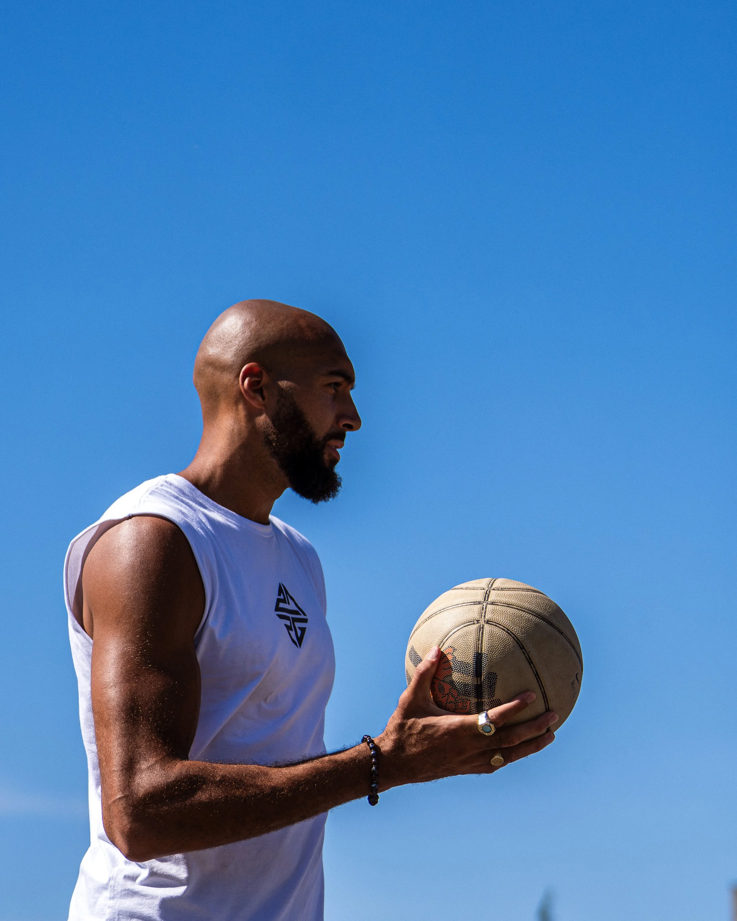 Un homme chauve avec une barbe tenant un ballon de basketball, portant un bracelet et une bague, contre un ciel bleu.