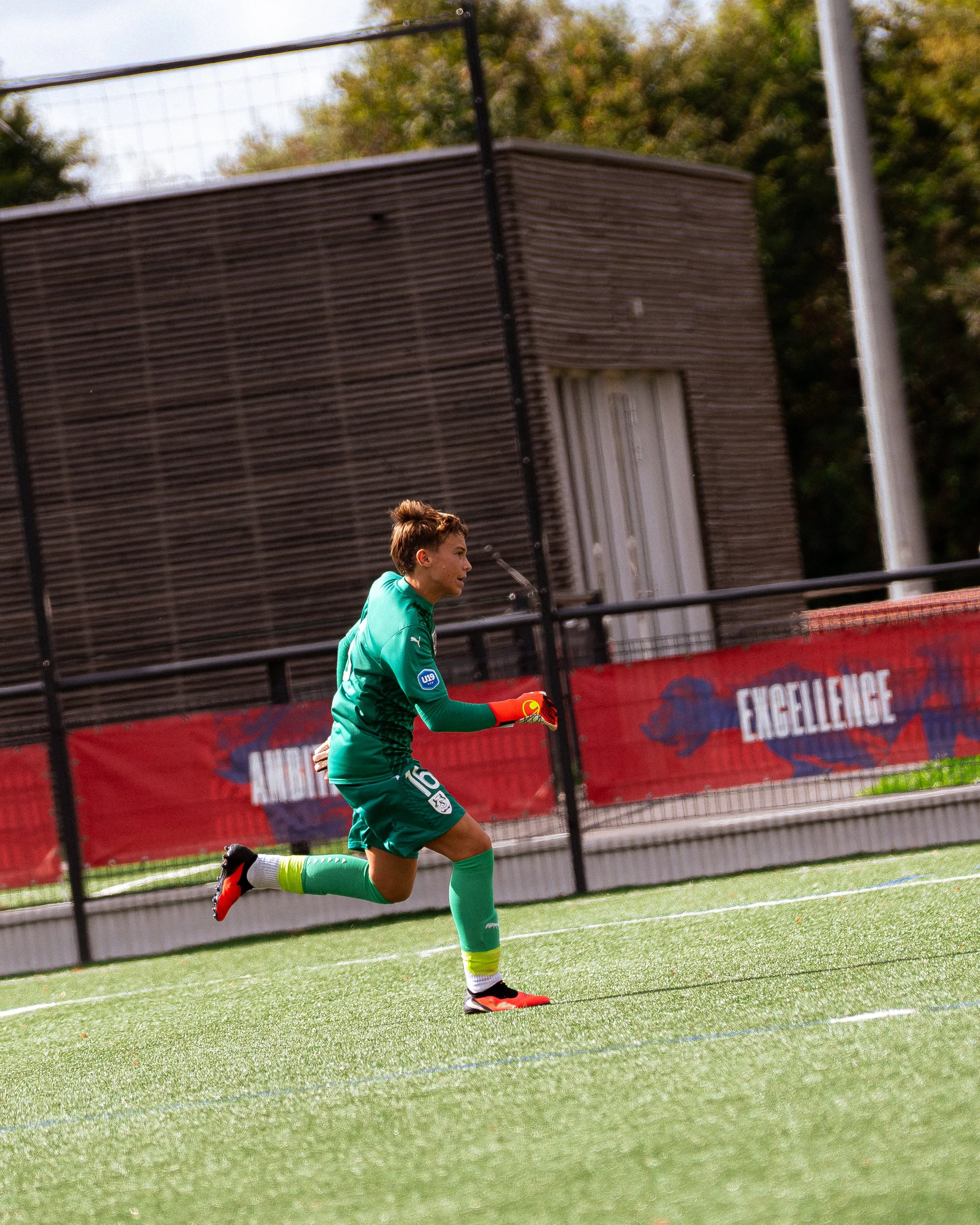 Un joueur de football en pleine course, vêtu d'un uniforme vert avec le numéro 16, sur un terrain de football avec des barrières et un bâtiment en arrière-plan.