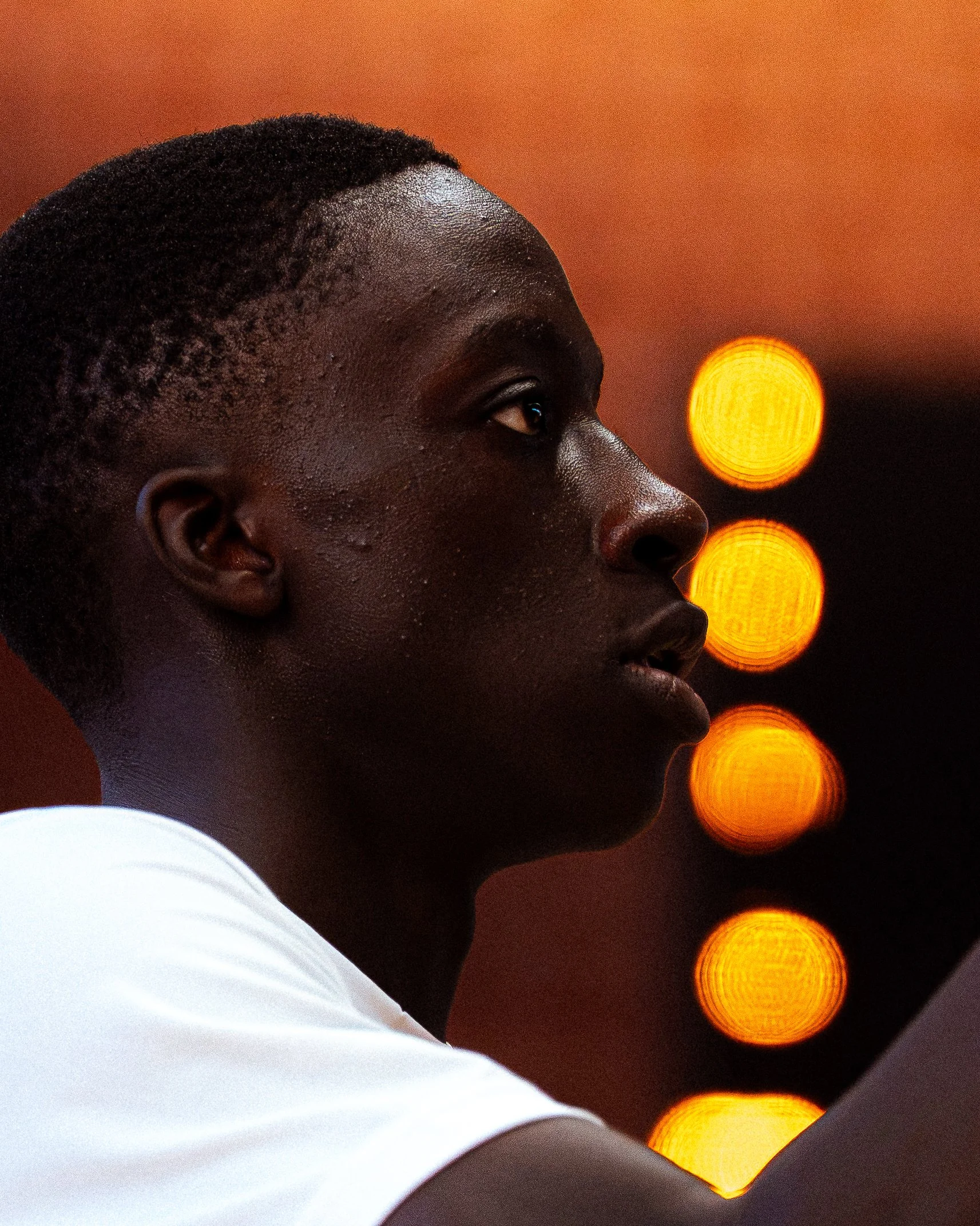 Portrait d'un jeune homme africain en profil, avec un fond flou de lumières orange. Il porte une chemise blanche.
