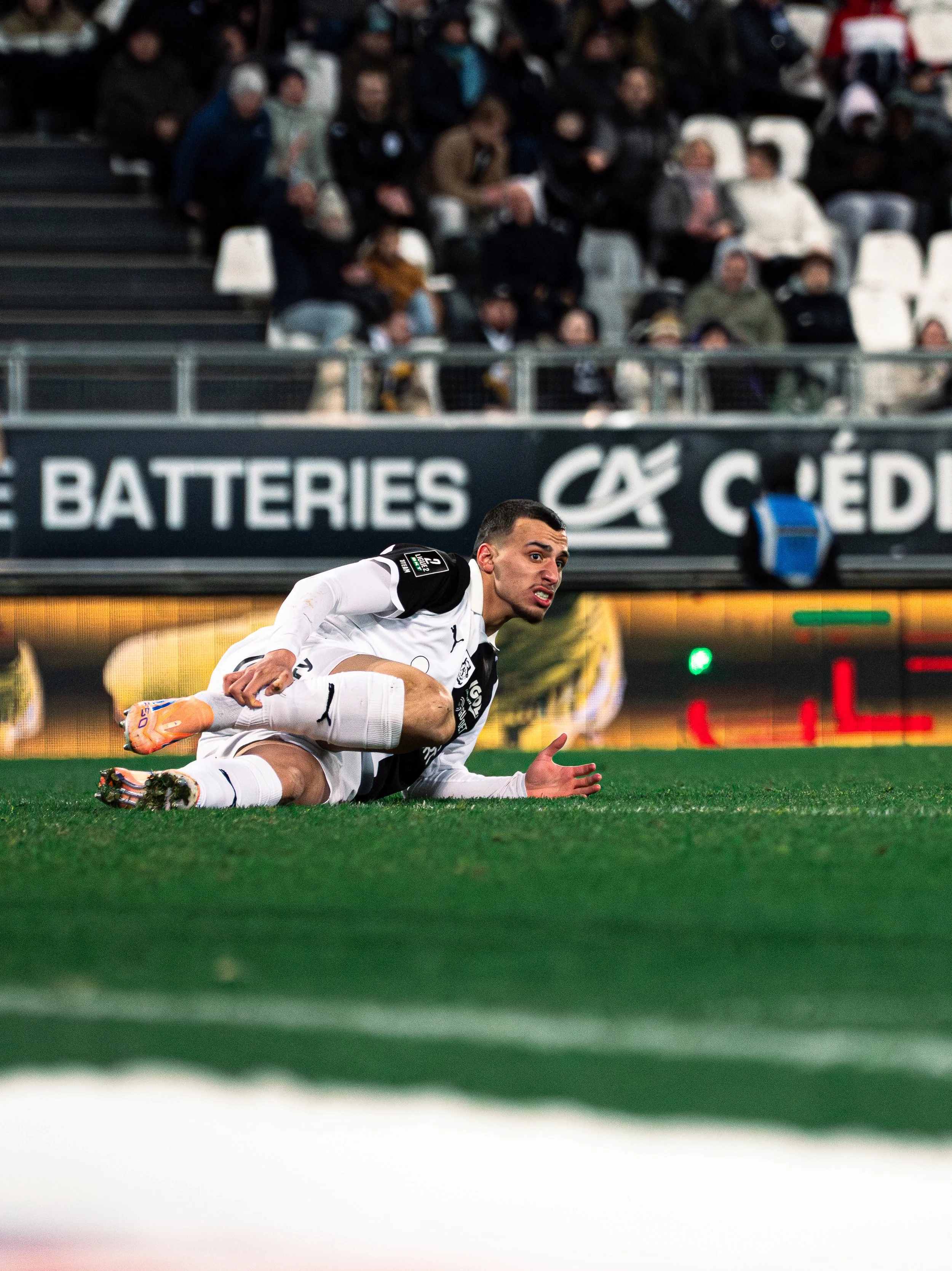 Un joueur de football en action, vêtu d'un maillot blanc, couché sur le terrain vert, avec un regard concentré, lors d'un match avec un public dans les gradins en arrière-plan.