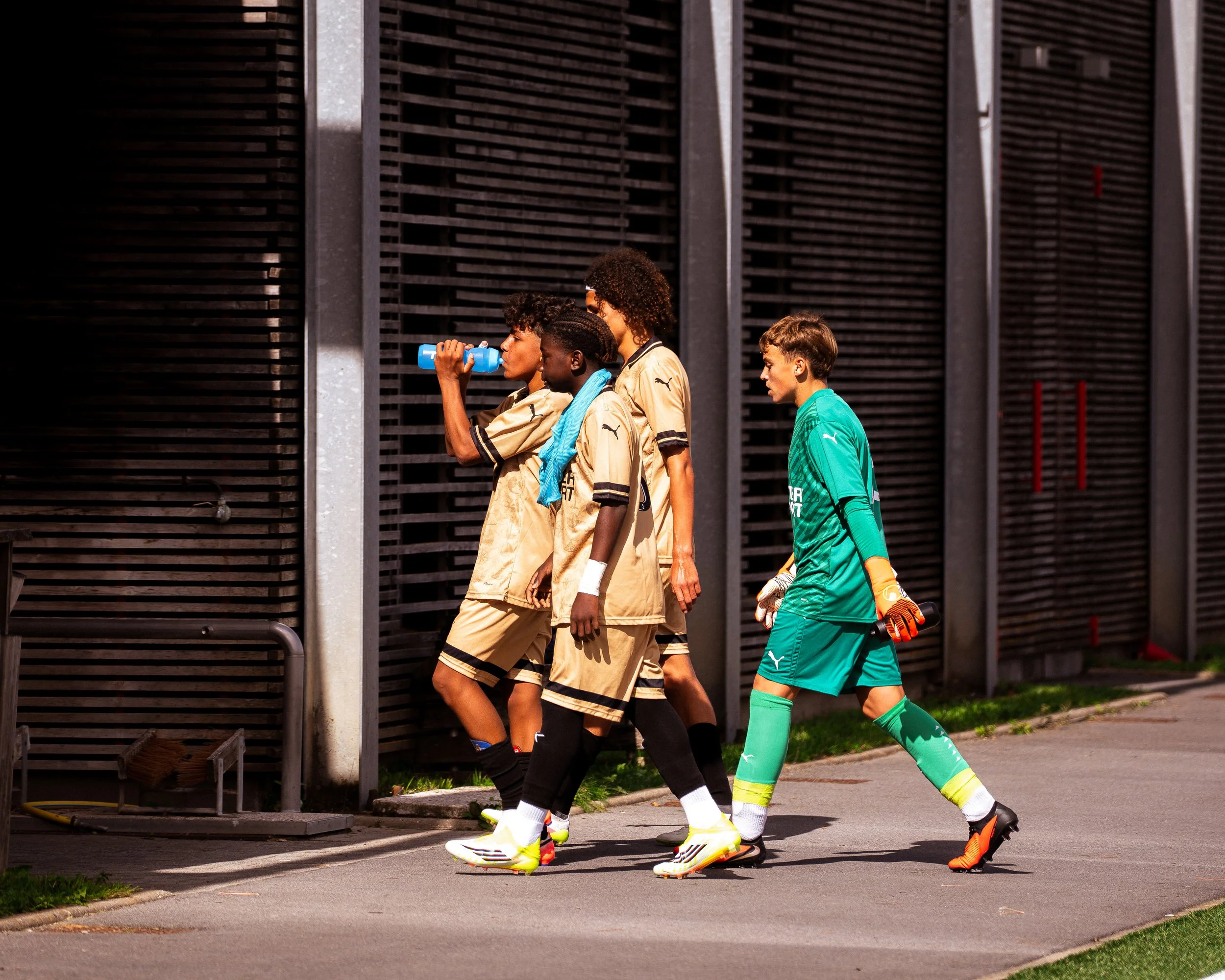Une équipe de jeunes footballeurs en tenue de sport, avec un gardien, marche en file devant une structure métallique, probablement avant ou après un match.