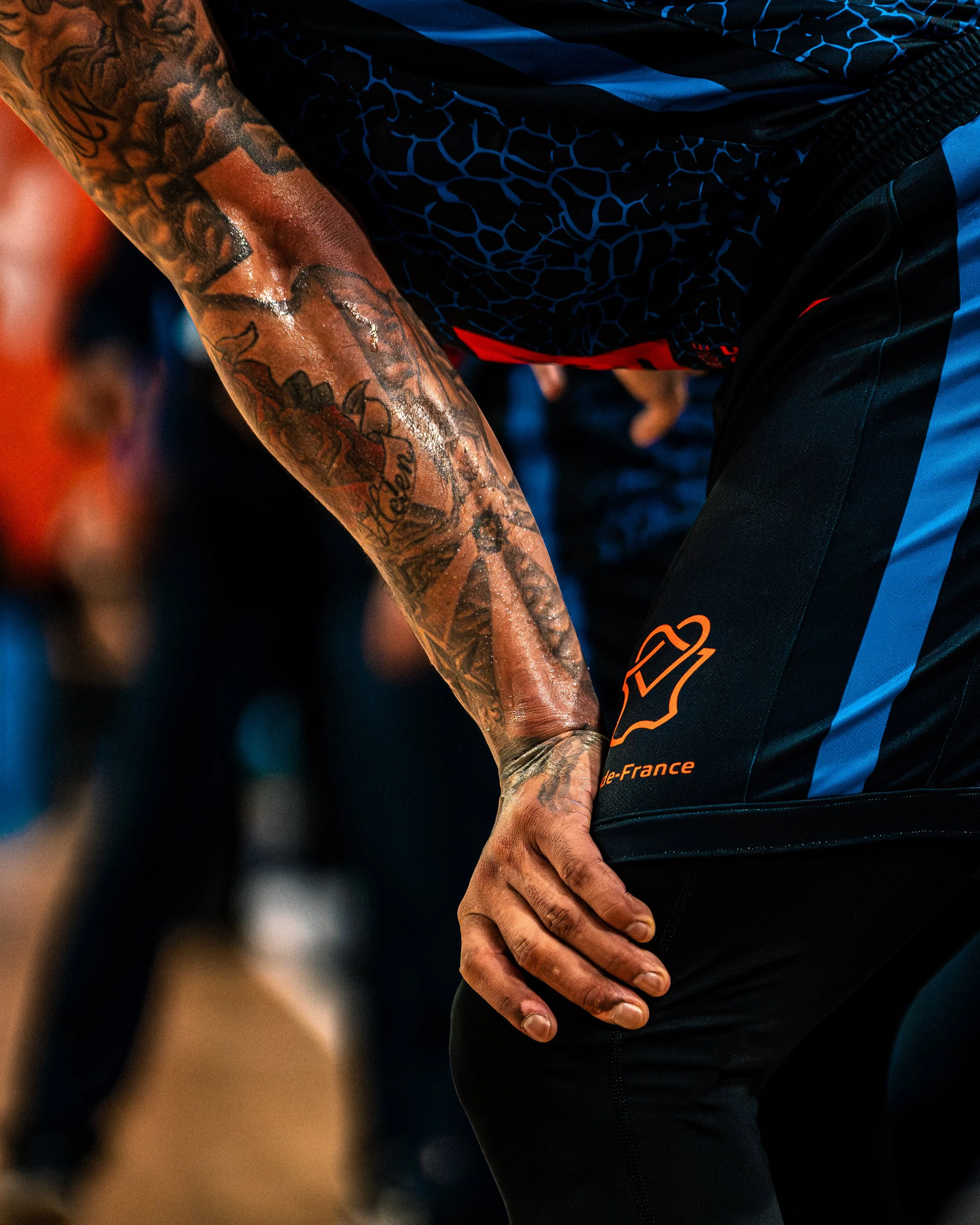 Bras droit tatoué, tenant la hanche avec une main, portant un vêtement sportif noir et bleu.