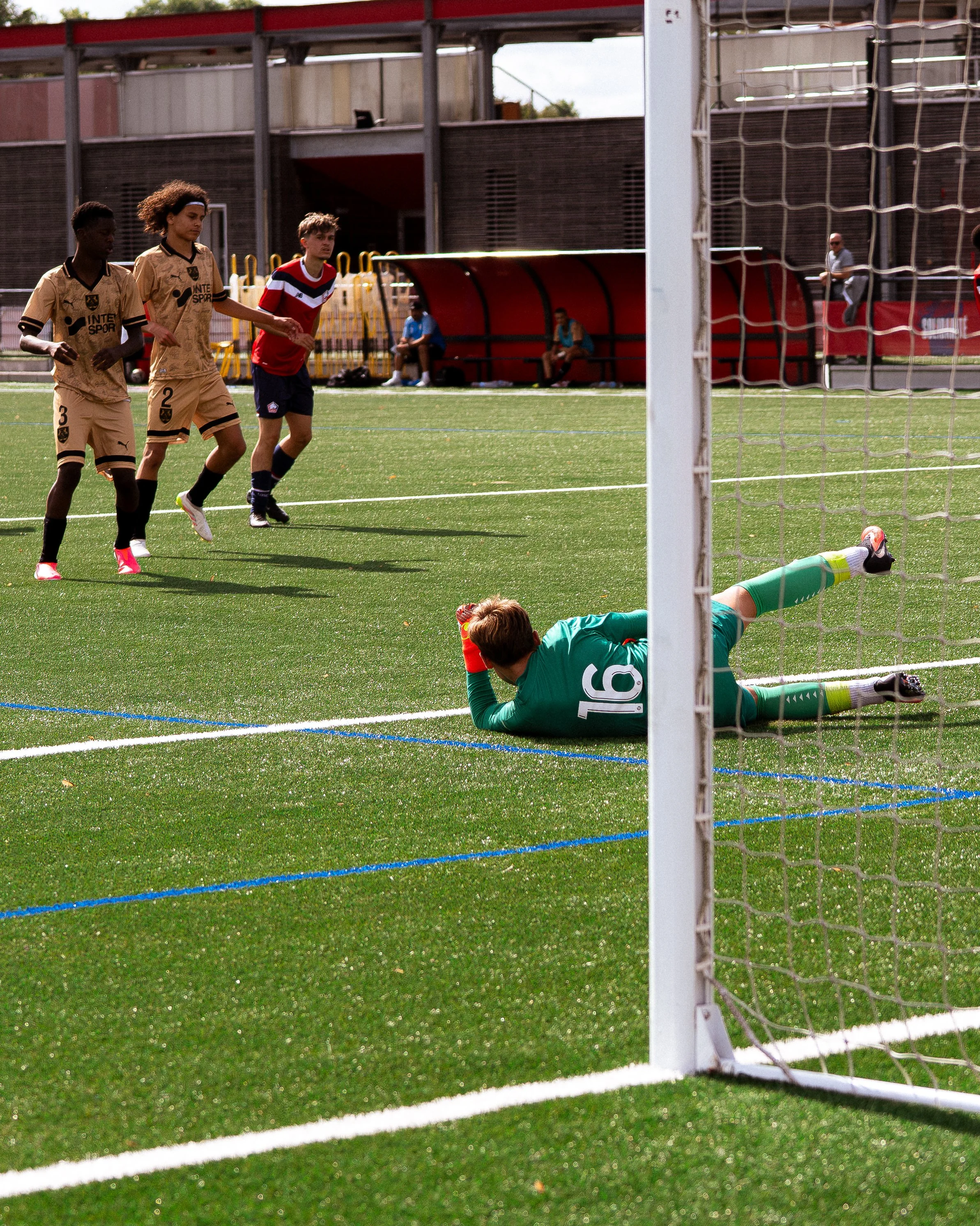 Une scène d'un match de football où un gardien est étendu au sol après avoir tenté d'arrêter un tir, tandis que deux joueuses de l'équipe adverse s'approchent, probablement après une tentative de but.