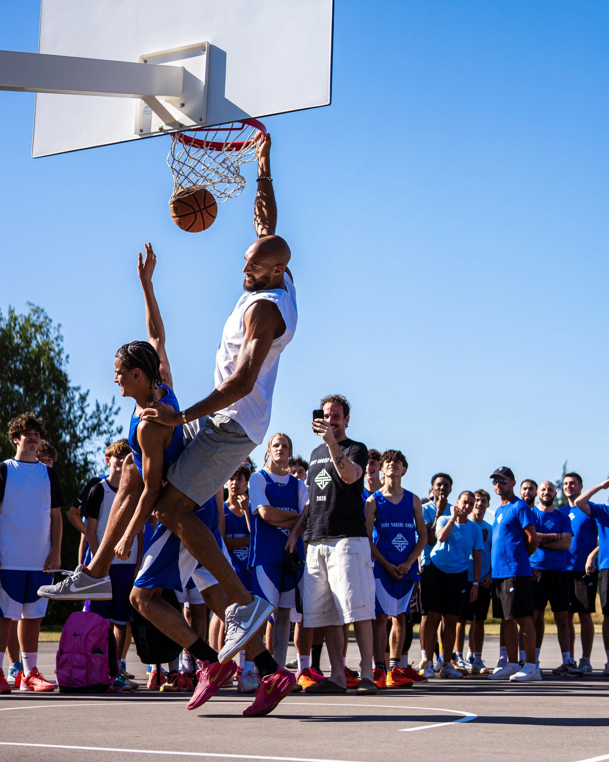 Un groupe de jeunes athlètes regarde un match de basketball sur un terrain extérieur, avec un joueur en action sautant pour tenter de faire un panier, sous un ciel clair et ensoleillé.