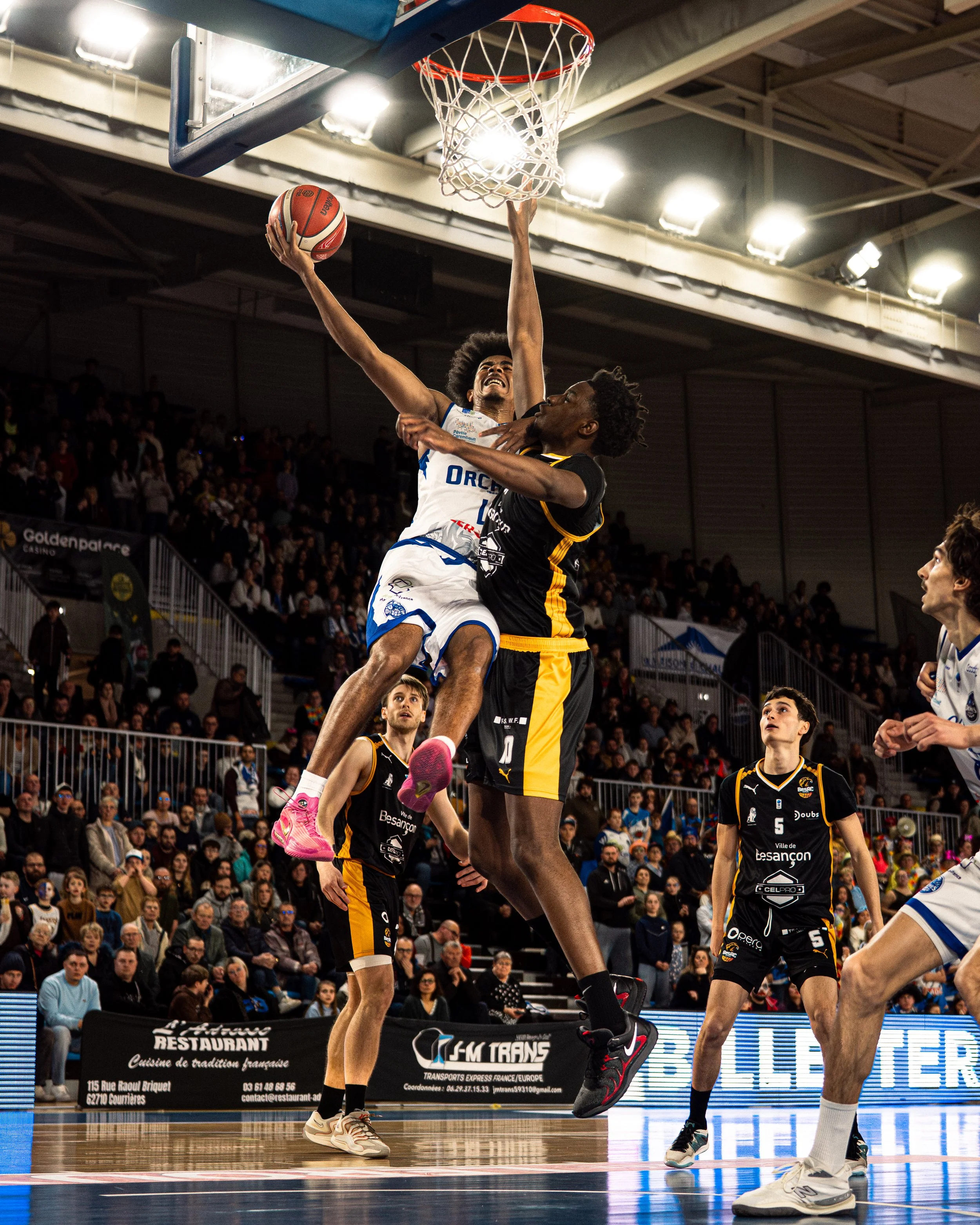 Deux joueurs de basketball, un en blanc et un en noir, en plein saut pour tenter une action au panier dans un match en intérieur, entourés d'un public nombreux.