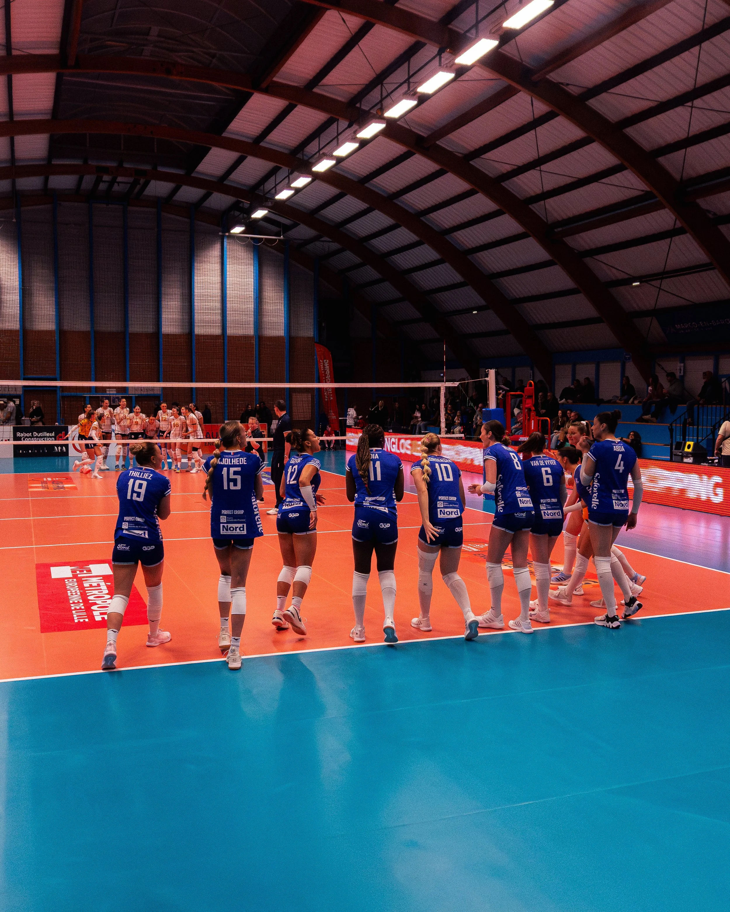 Une photo de l'intérieur d'une salle de volleyball où une équipe féminine en uniforme bleu est en train de se préparer pour jouer, avec d'autres personnes dans le fond. La salle a un plafond en arches en bois et un sol en vinyle orange avec une ligne