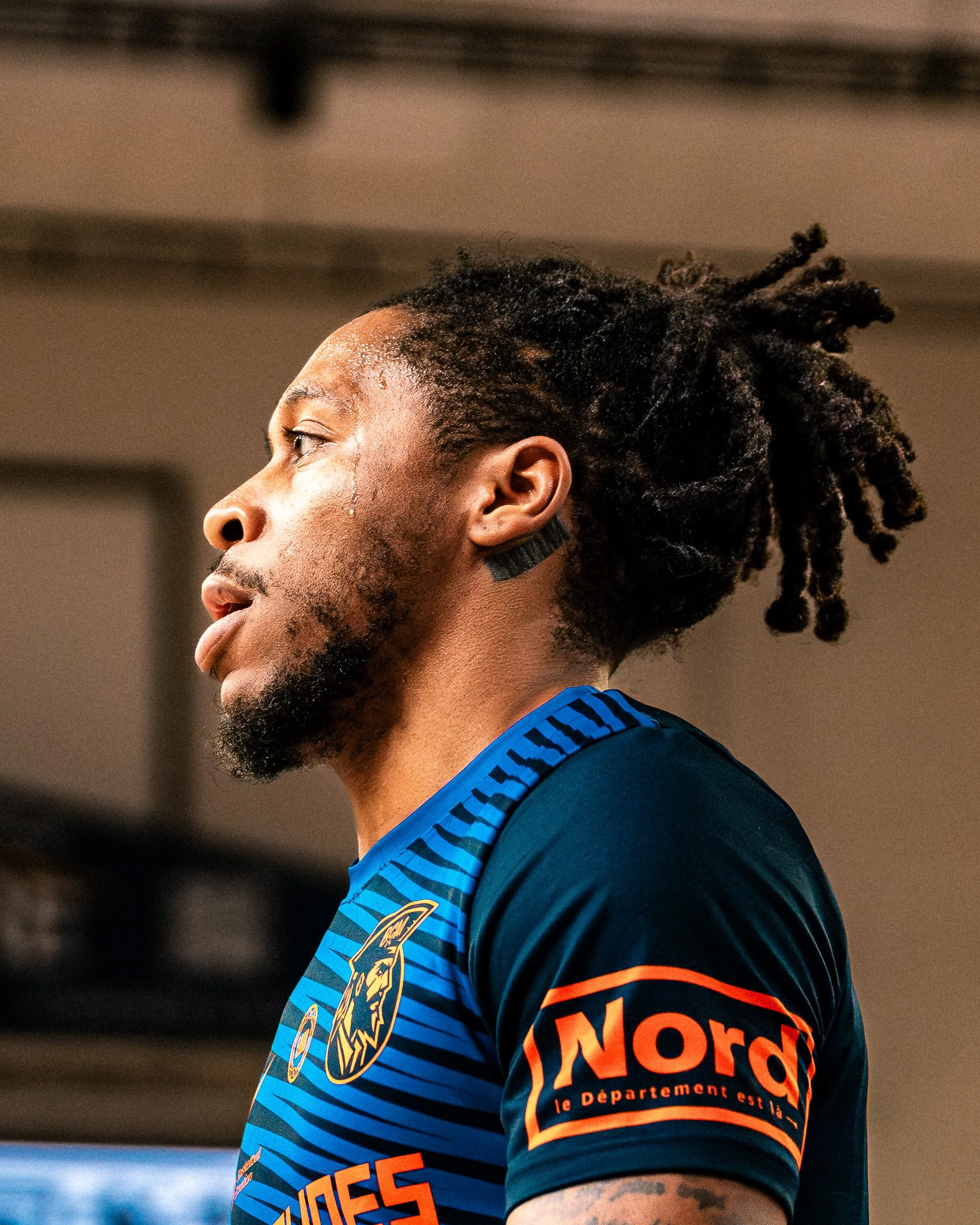 Vue de profil d'un homme avec des dreadlocks, portant un maillot de sport bleu avec des rayures noires et des logos, dans un environnement intérieur.