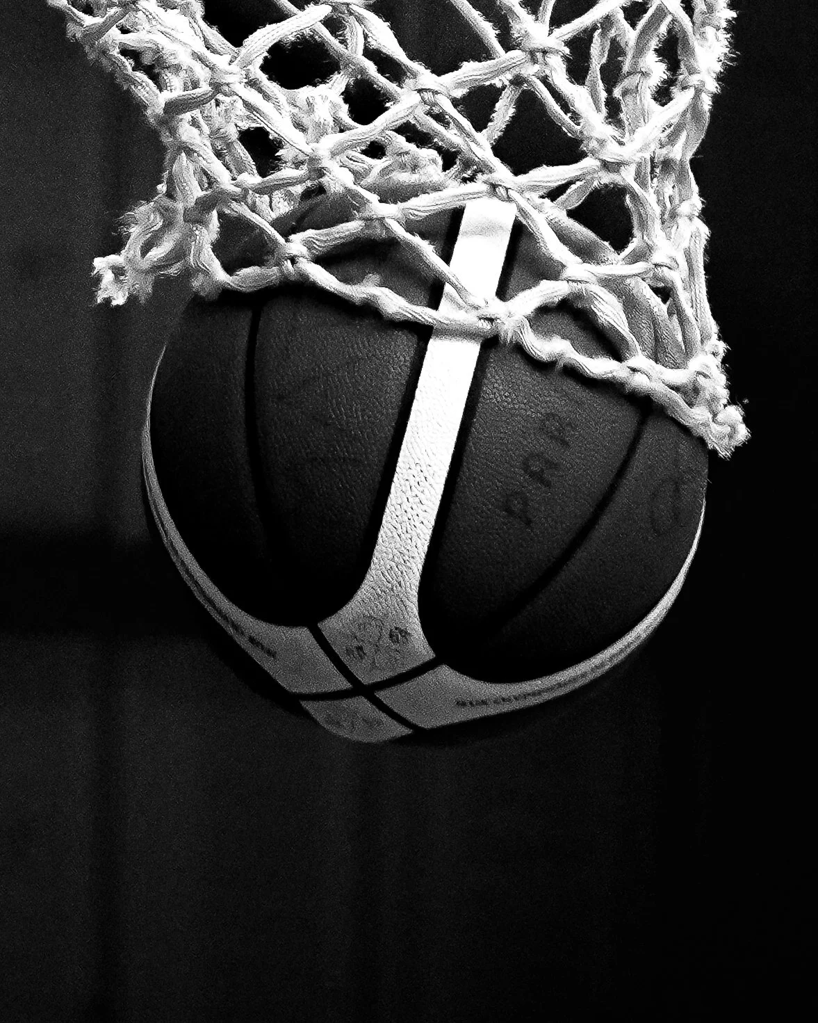Une balle de basket suspendue dans un panier de basketball, vue de face, en noir et blanc.