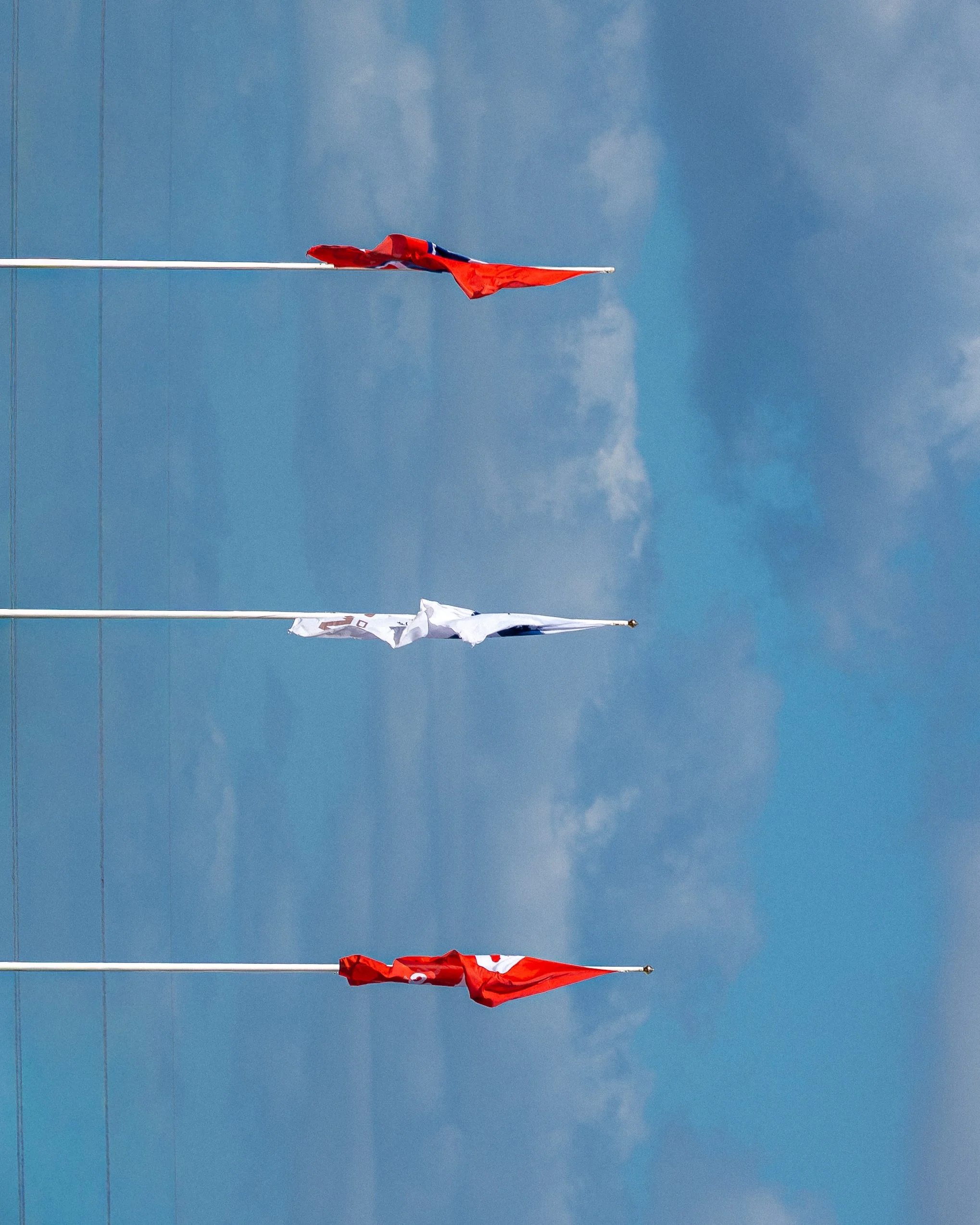 Deux drapeaux, un rouge avec une partie blanche et un autre blanc, flottent sur des mâts sous un ciel bleu avec quelques nuages.