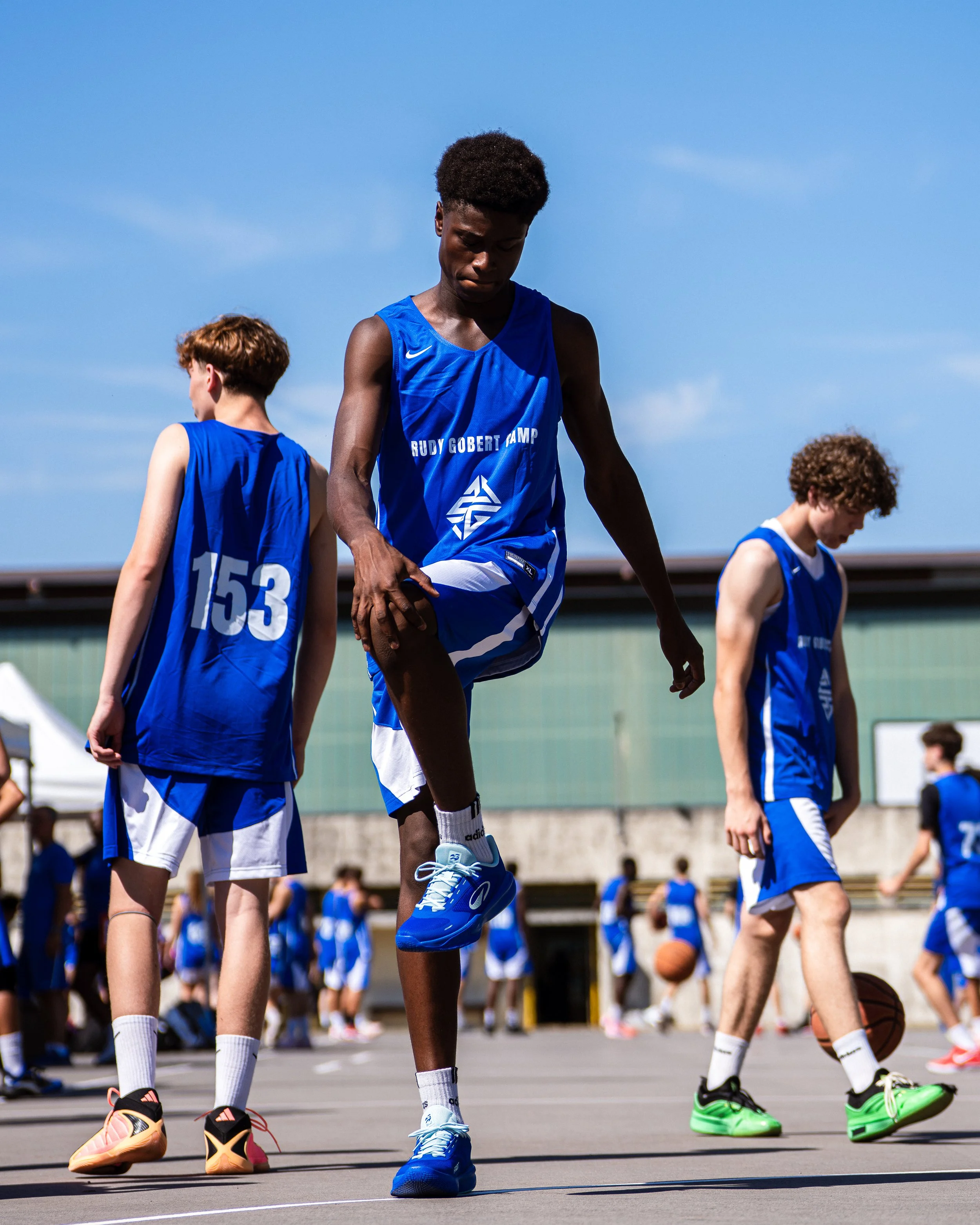 Des jeunes athlètes en tenue de sport bleue s'échauffent ou jouent sur un terrain de basketball extérieur par une journée ensoleillée, avec un bâtiment en arrière-plan.
