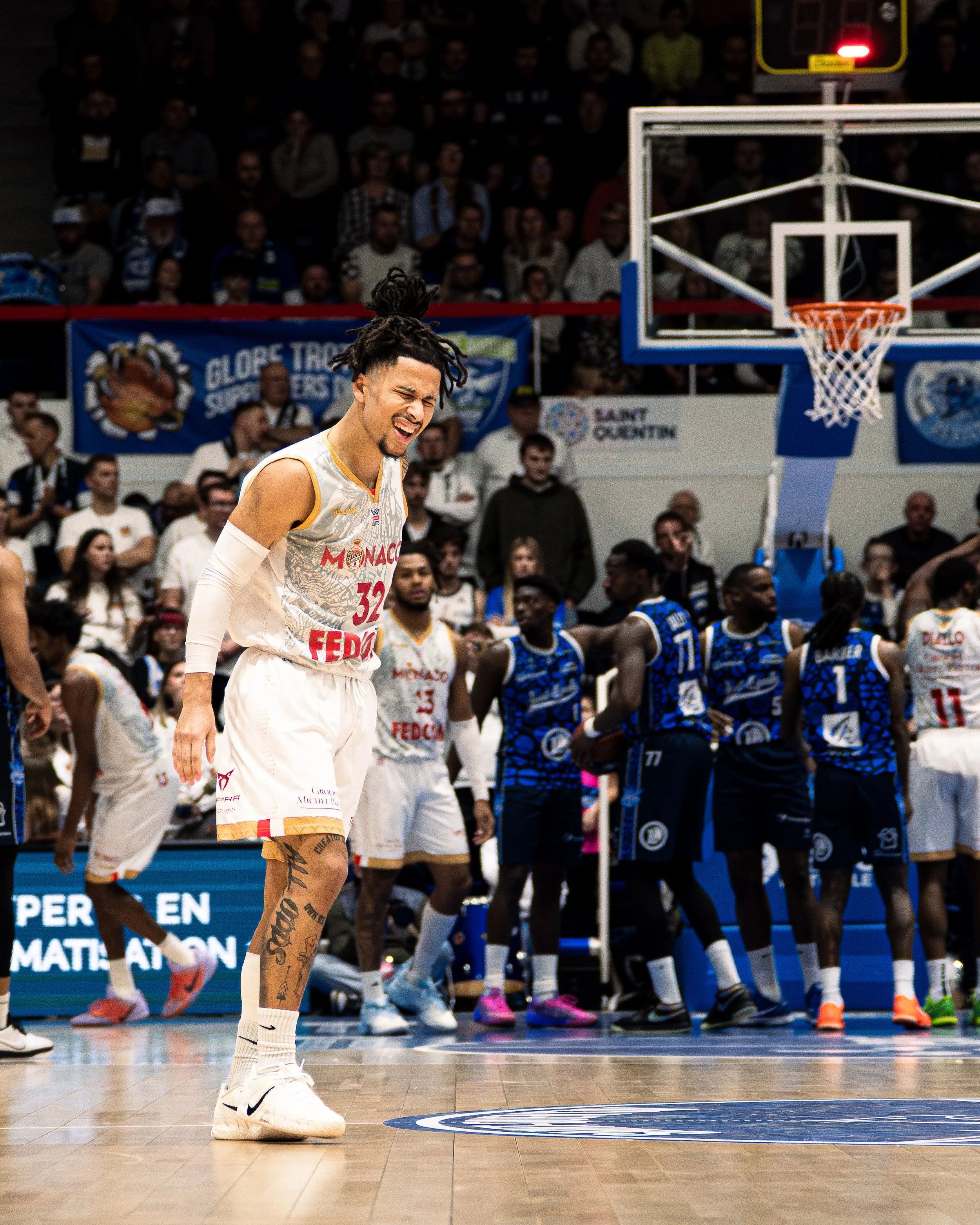 Un joueur de basketball en tenue blanche avec le logo Fedcom, plein de tatouages sur la jambe, semble souffrir ou être déçu, lors d'un match, avec des spectateurs et d'autres joueurs en fond.