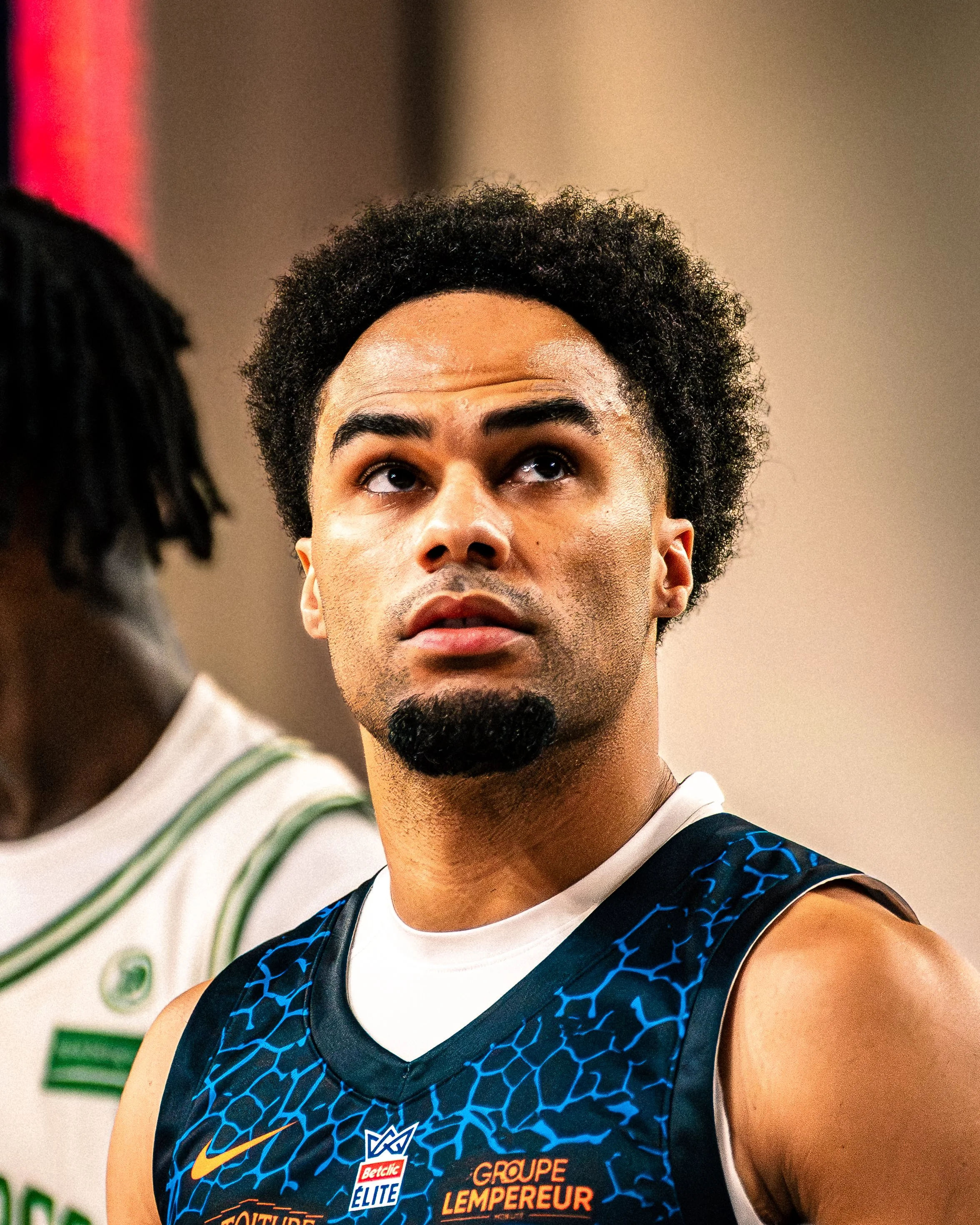 Un homme portant un maillot de basketball avec plusieurs logos, regardant vers le haut, dans un environnement intérieur.