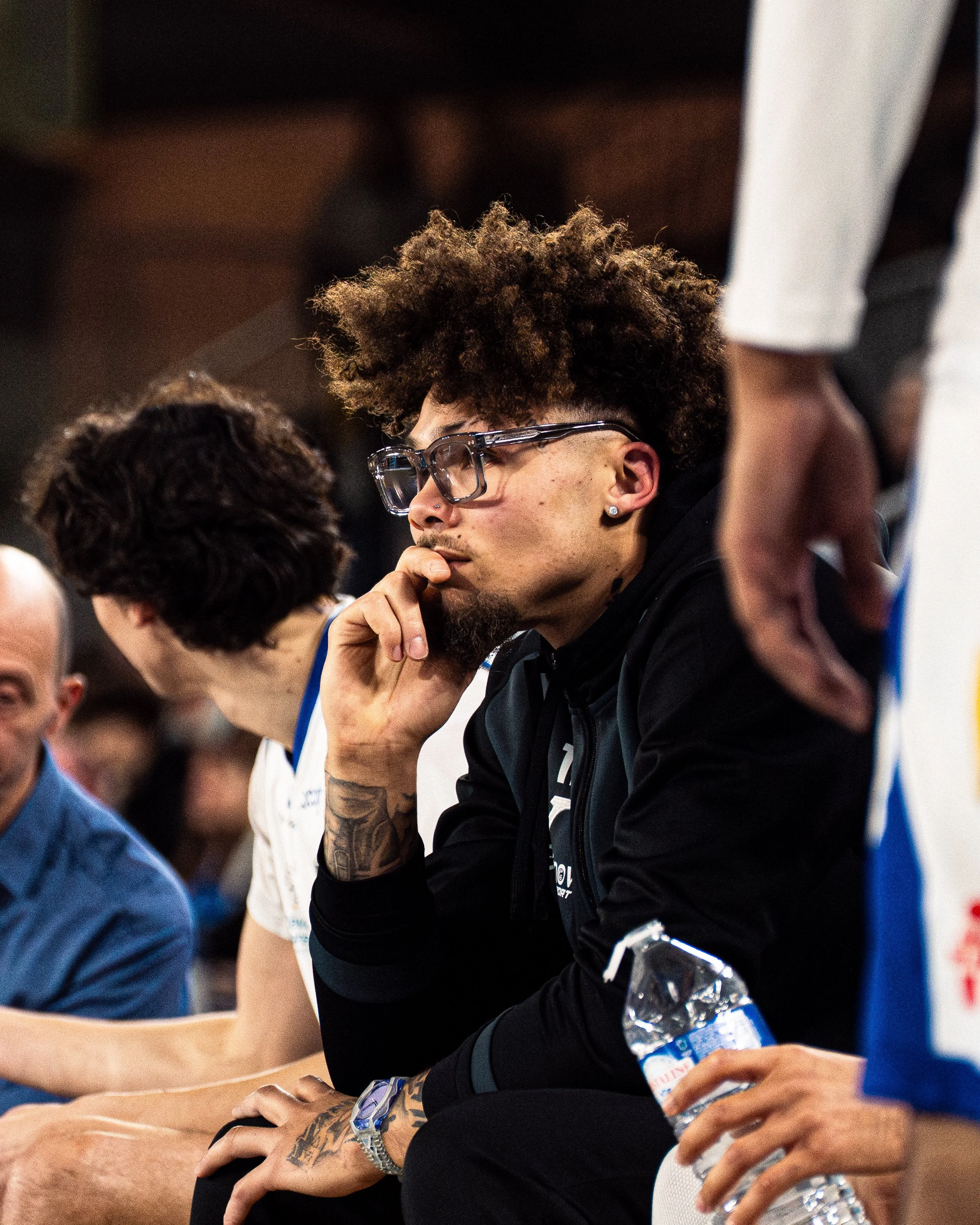 Un jeune homme portant des lunettes, assis à une table, semble concentré ou pensif. Il a une barbe et une peau claire. Il porte un sweat noir avec des détails blancs. Il tient une bouteille d'eau en plastique dans sa main droite. La scène semble se d