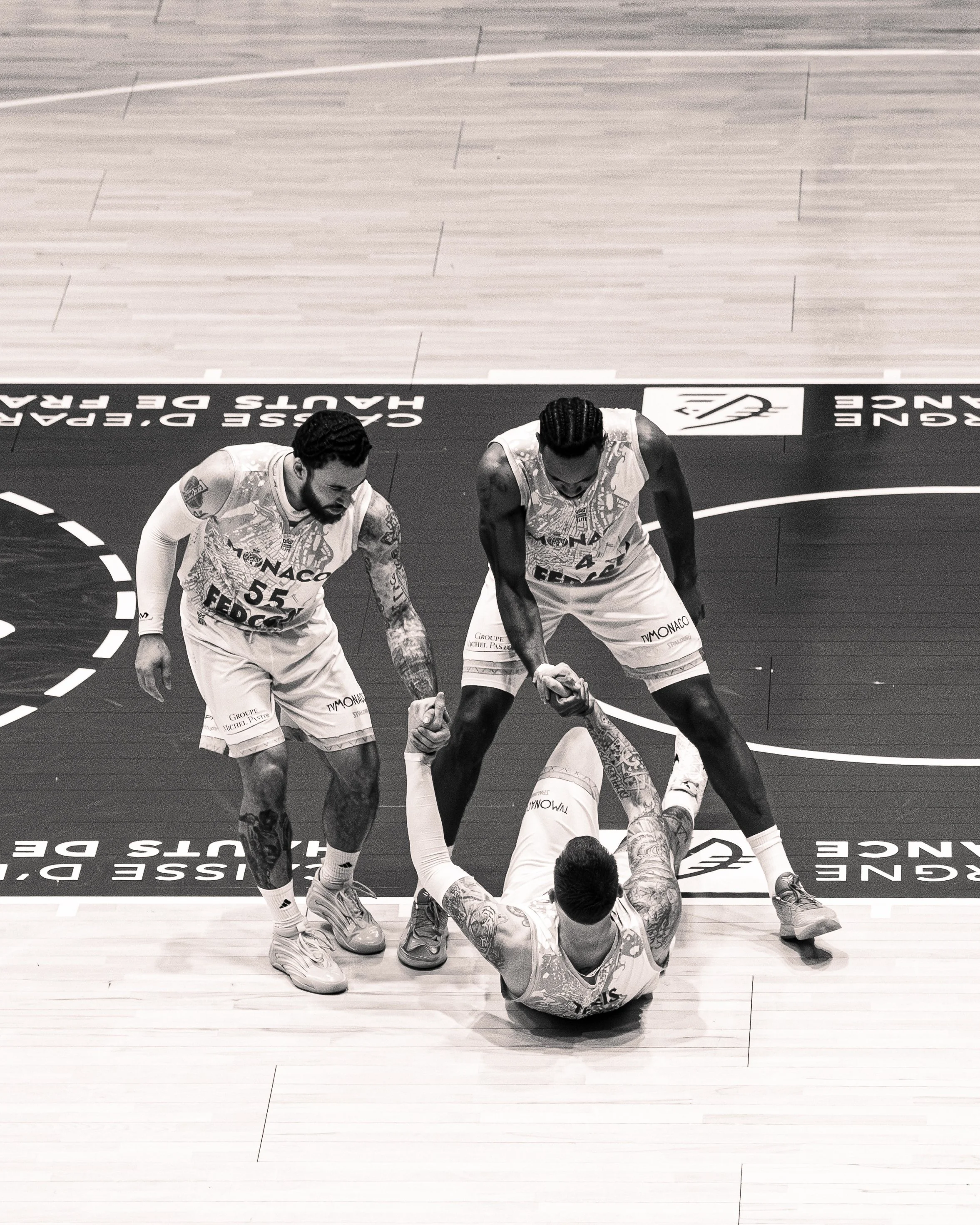 Deux joueurs de basketball en maillots avant un match, aidant une personne à se relever sur un terrain en bois. La scène se déroule dans une salle de sport. Leurs maillots portent le nom de Monaco et l'inscription 'Groupe Michel Pastore'.