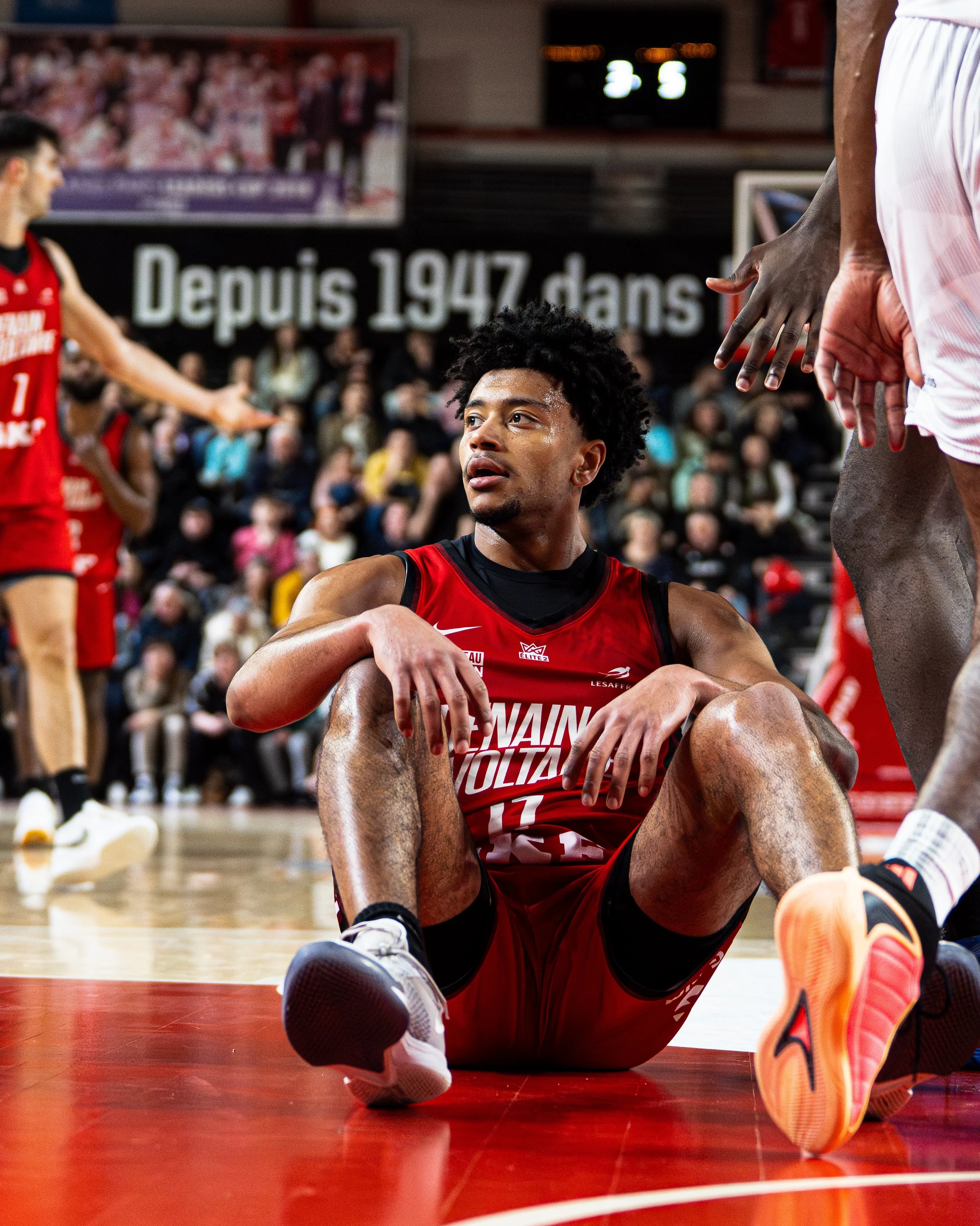 Un joueur de basketball allongé sur le sol lors d'un match, portant un maillot rouge avec le numéro 11, en train de se faire soigner ou de se reposer, avec d'autres joueurs et la foule en arrière-plan, et un tableau avec une annonce en français 'Depu