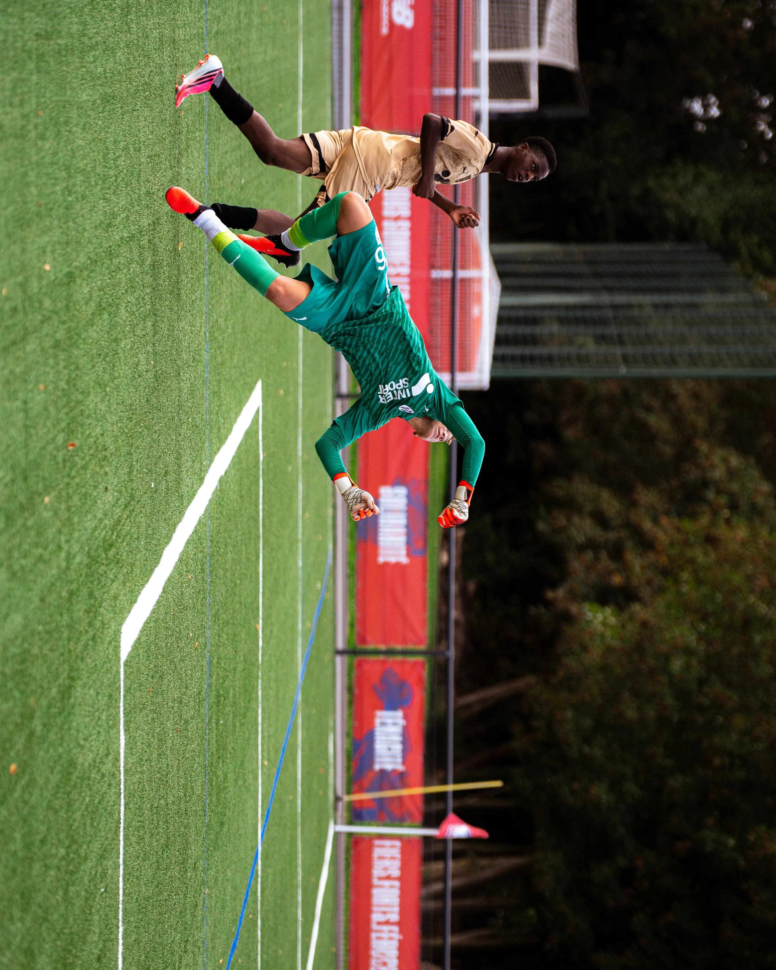 Juvenile soccer match on a green field, player in a green jersey leaning forward, opponent in a beige jersey running, stadium in the background.