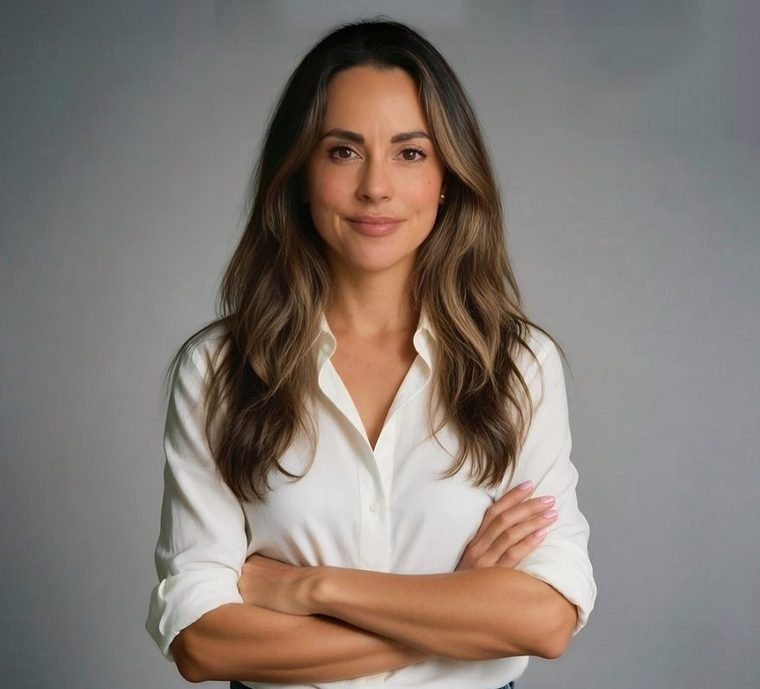 A woman with long wavy brown hair and light skin, wearing a white collared shirt, standing with her arms crossed in front of a gray background, looking confidently at the camera.