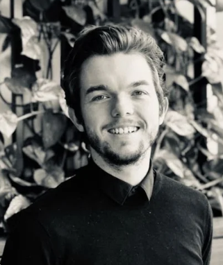 Young man with a beard and mustache smiling, standing outdoors with foliage in the background.