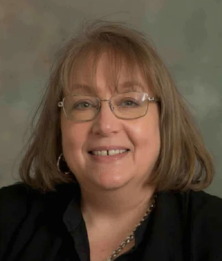 A woman with shoulder-length light brown hair, wearing glasses, a black top, and a silver necklace, smiling at the camera.