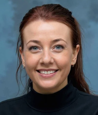 Portrait of a woman with auburn hair, blue eyes, wearing a black turtleneck, smiling against a blue background.