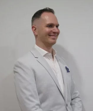 Man in a light gray suit and white shirt smiling against a plain gray background.