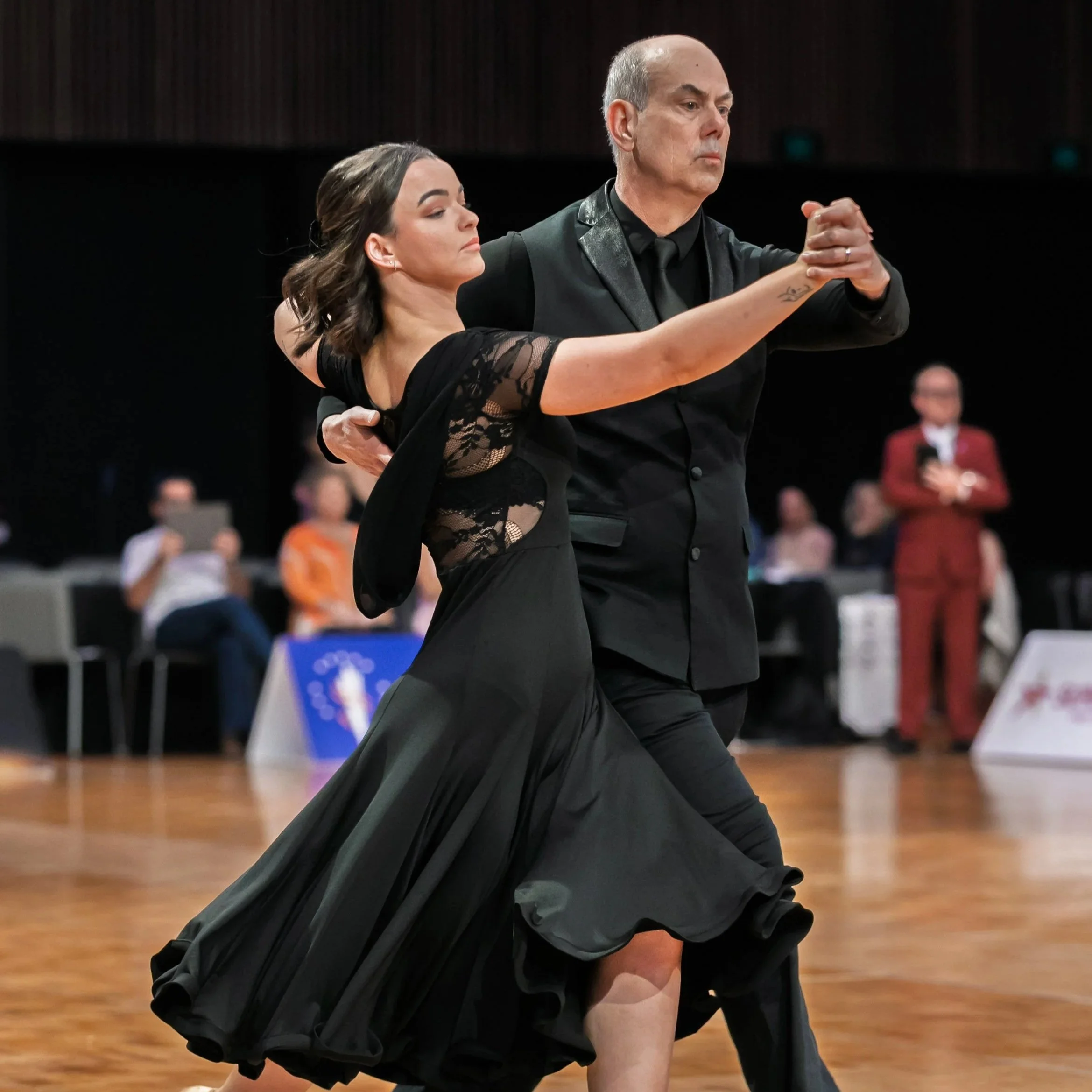 A middle-aged man and woman dressed in formal dance attire, dancing in a ballroom. The woman wears a black dress with lace details, and the man wears a black tuxedo. They are holding each other and dancing closely, with focused expressions.
