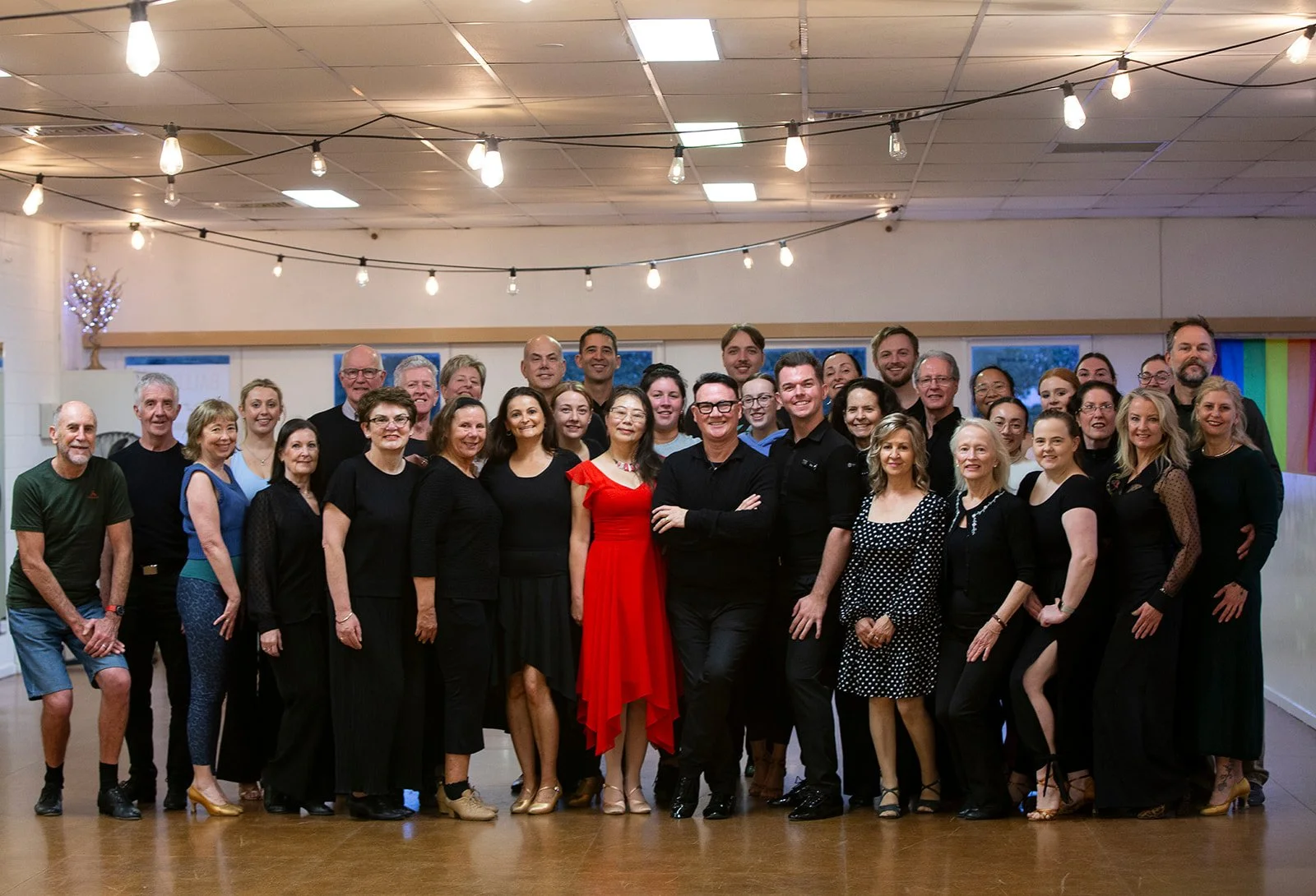 A large group of diverse people posing together indoors for a photo, with some smiling and wearing casual to semi-formal attire, in a well-lit room with string lights and windows in the background.