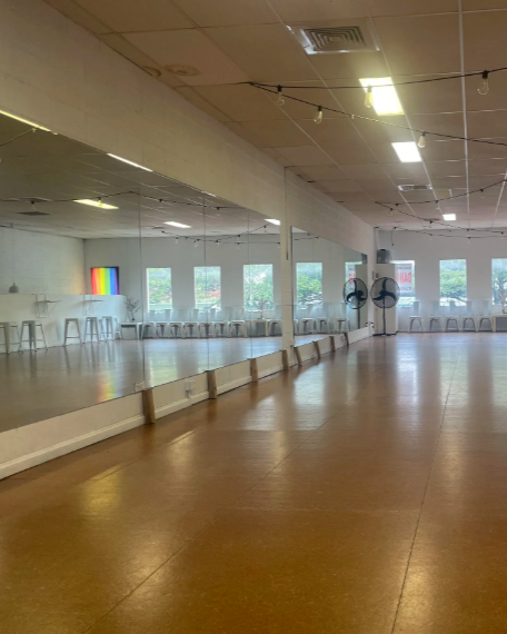 Empty dance studio with large mirror, wooden floor, and chairs along the walls, decorated with colorful artwork and windows letting in natural light.