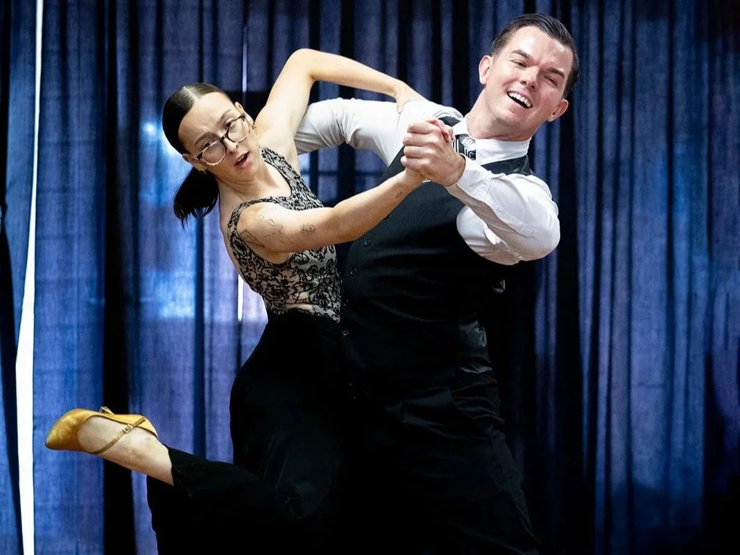 A man and woman are dancing together indoors, the man is smiling and wearing a tuxedo with a white shirt, and the woman is wearing glasses, a black lace dress, and yellow shoes, with her hair tied back.