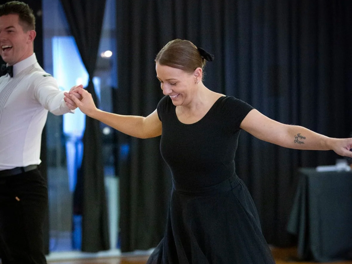 A woman with a tattoo on her right arm and hair tied back is dancing with a man in a tuxedo and bow tie, both holding hands in a dance hall.
