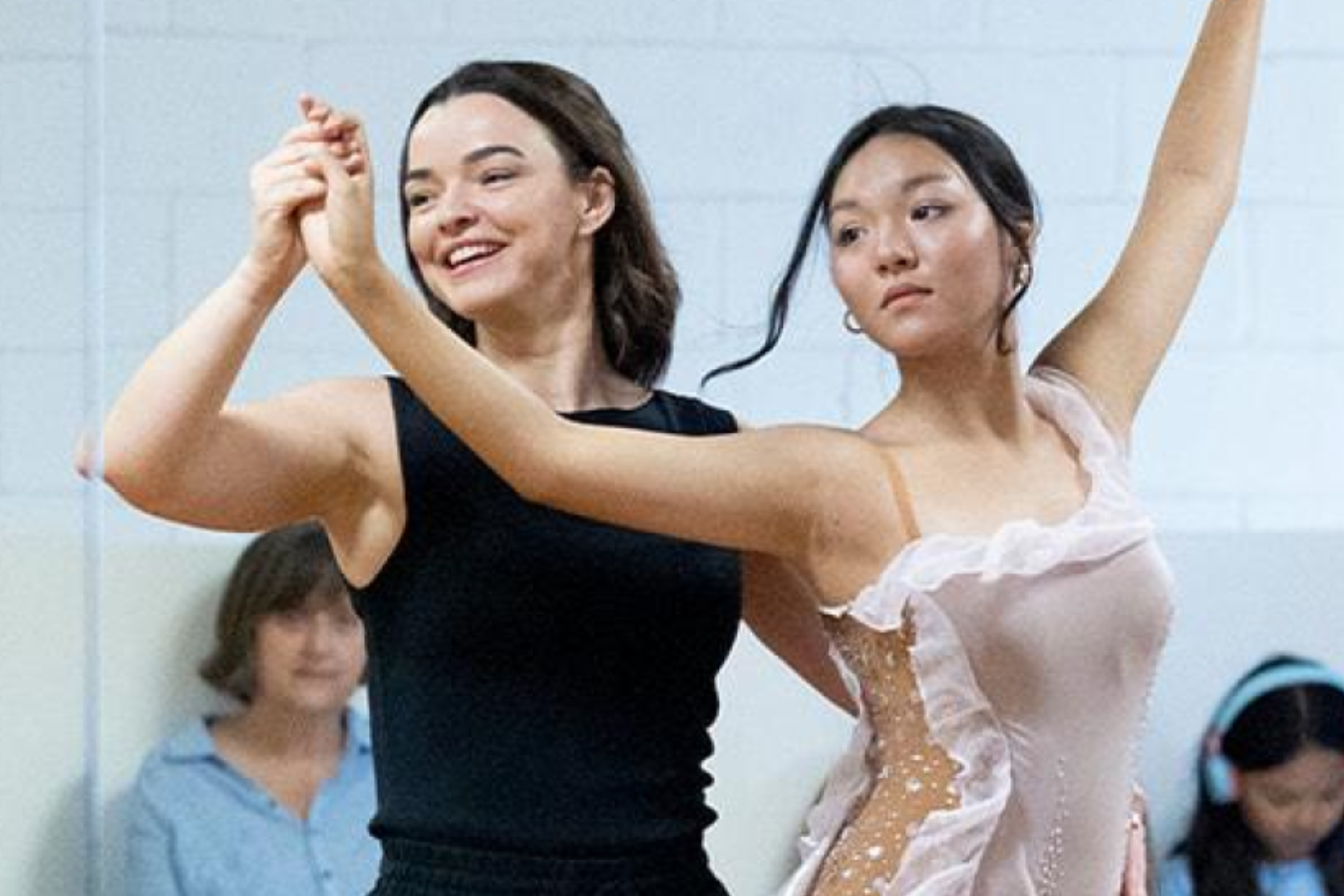 Two women are dancing and holding hands, one wearing a black dress and the other wearing a light pink ballet costume. They are in a room with a white background, and there are seated spectators watching.