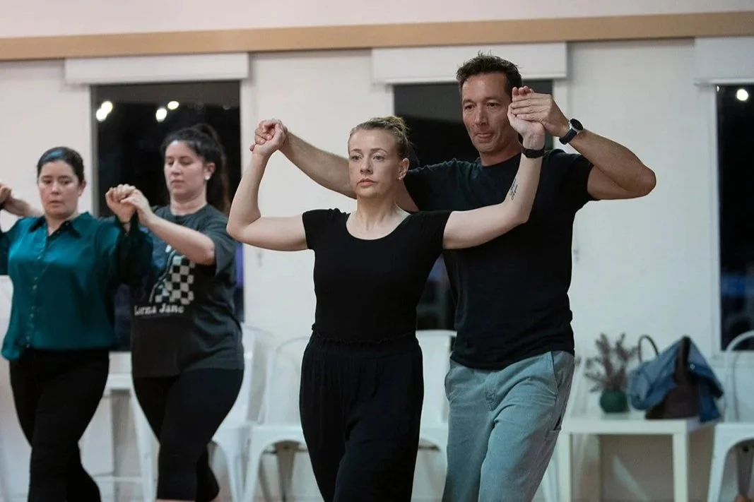 Group of people practicing dance or exercise in a studio, focusing on a young woman with blonde hair and a man, both with serious expressions, holding hands in the foreground.