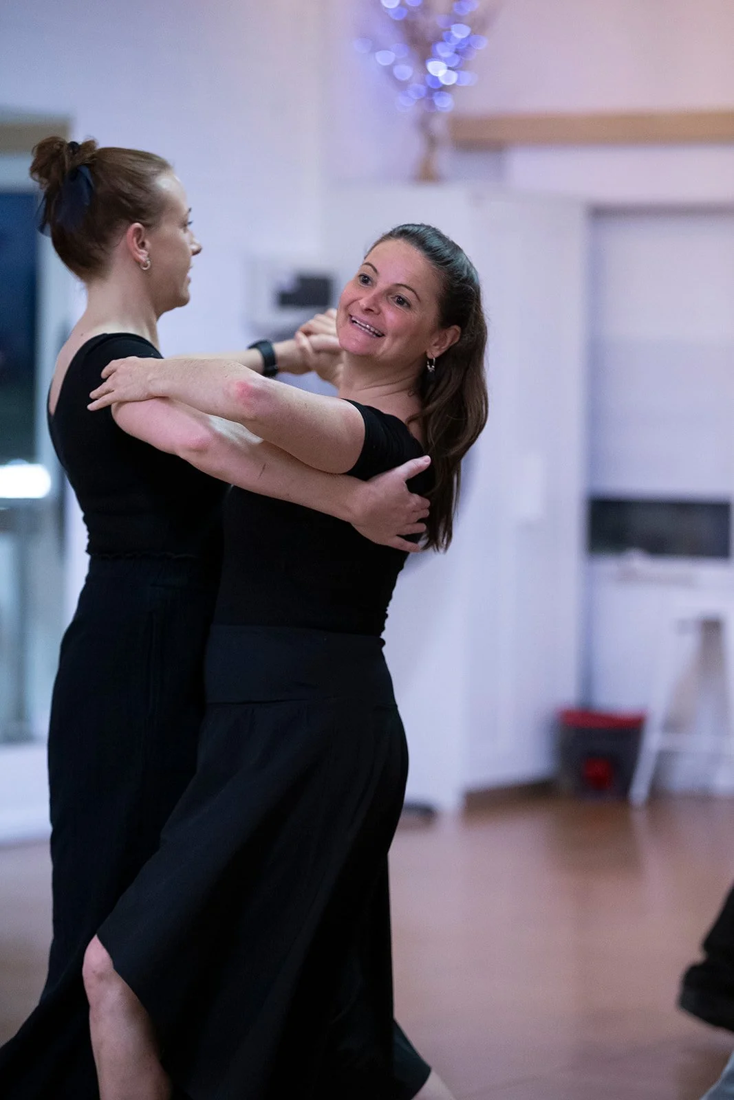 Two women with black clothing dancing closely, smiling, with a blurred festive background.