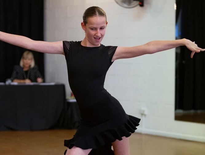 A young girl in a black dance outfit performing a dance pose in a studio.