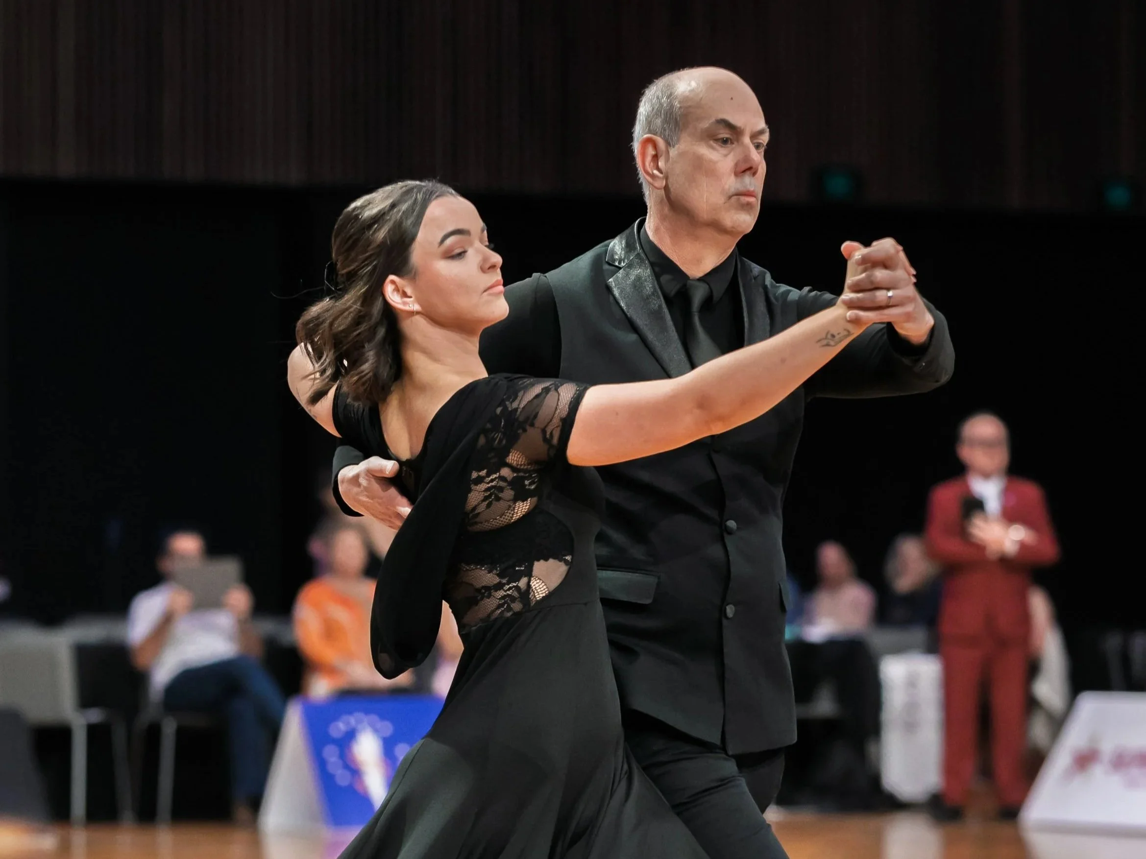 A man and woman participating in a ballroom dance competition, dressed in formal dance attire, focused and in motion on the dance floor, with spectators blurred in the background.