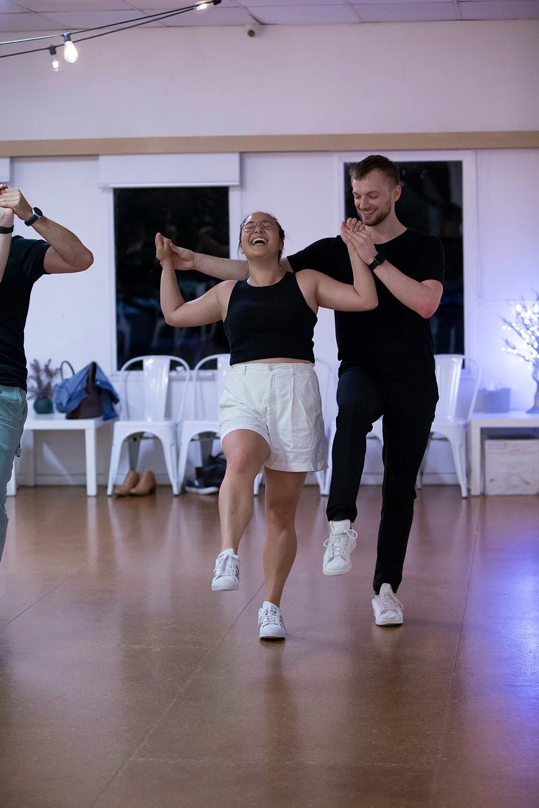 Two people dancing happily in a room with wooden floors, white walls, and string lights overhead. The woman wears a black tank top and white shorts, and the man wears a black t-shirt and black pants. They hold hands, smiling and raising their legs in a dance move.