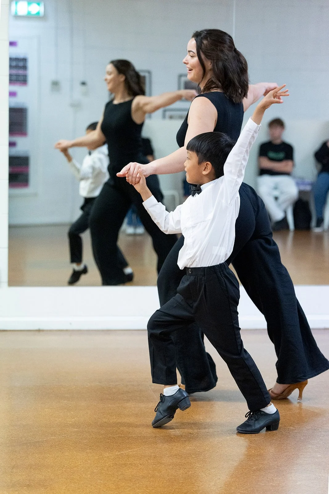 A woman and a boy dance together in a studio, with both smiling. The woman is in a black dress, and the boy is in white shirt and black trousers. Their reflection is visible in a mirror behind them, with other dancers in the background.