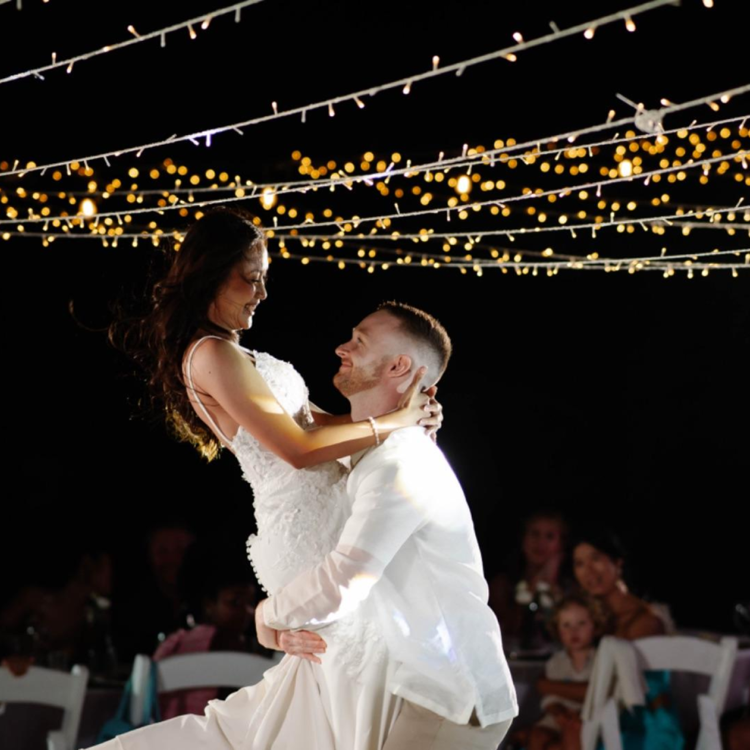 A bride and groom dancing at their wedding reception under string lights, with guests watching in the background.