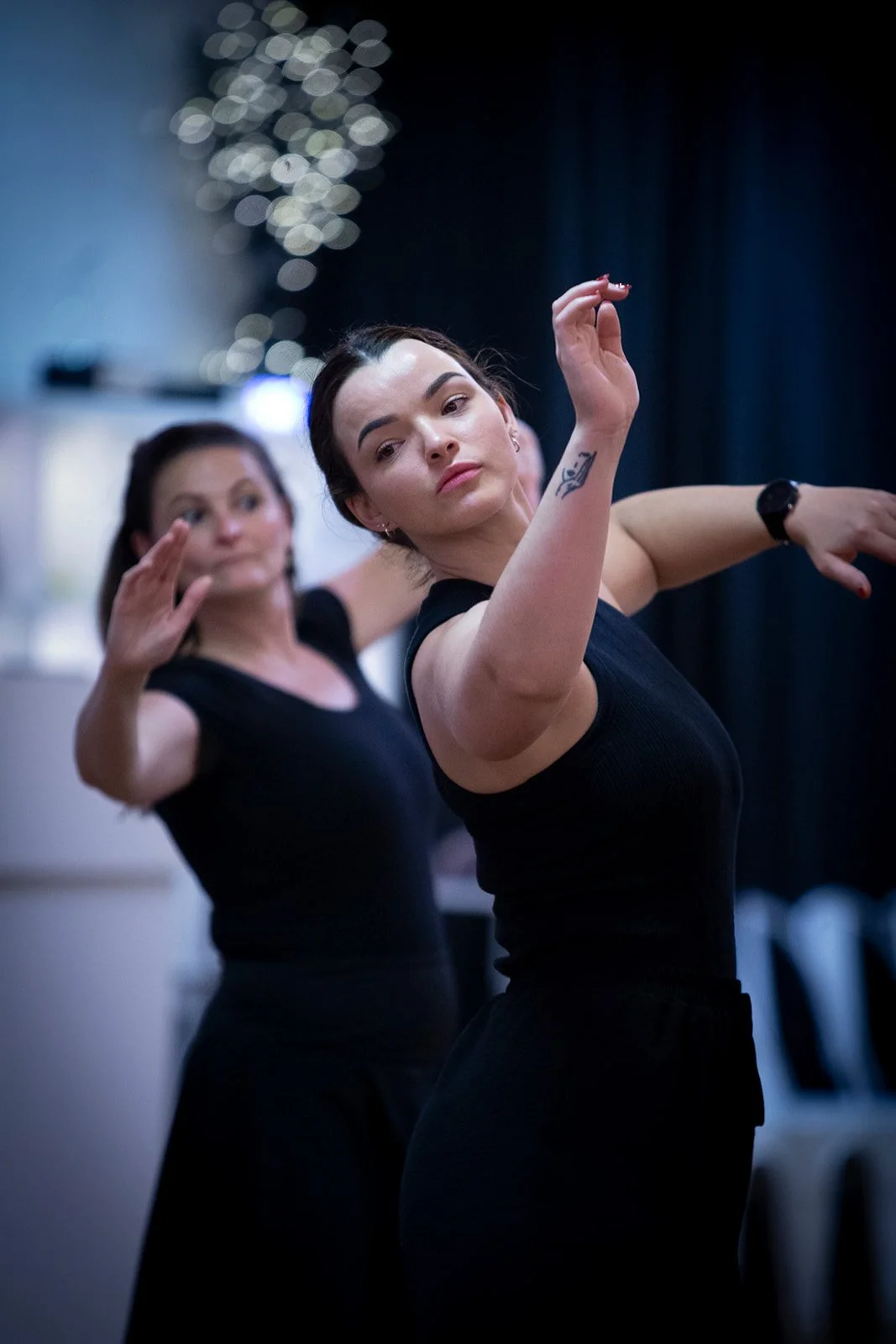 Two women practicing dance moves in a dimly lit studio, wearing black outfits, with a blurred background and decorative lights.