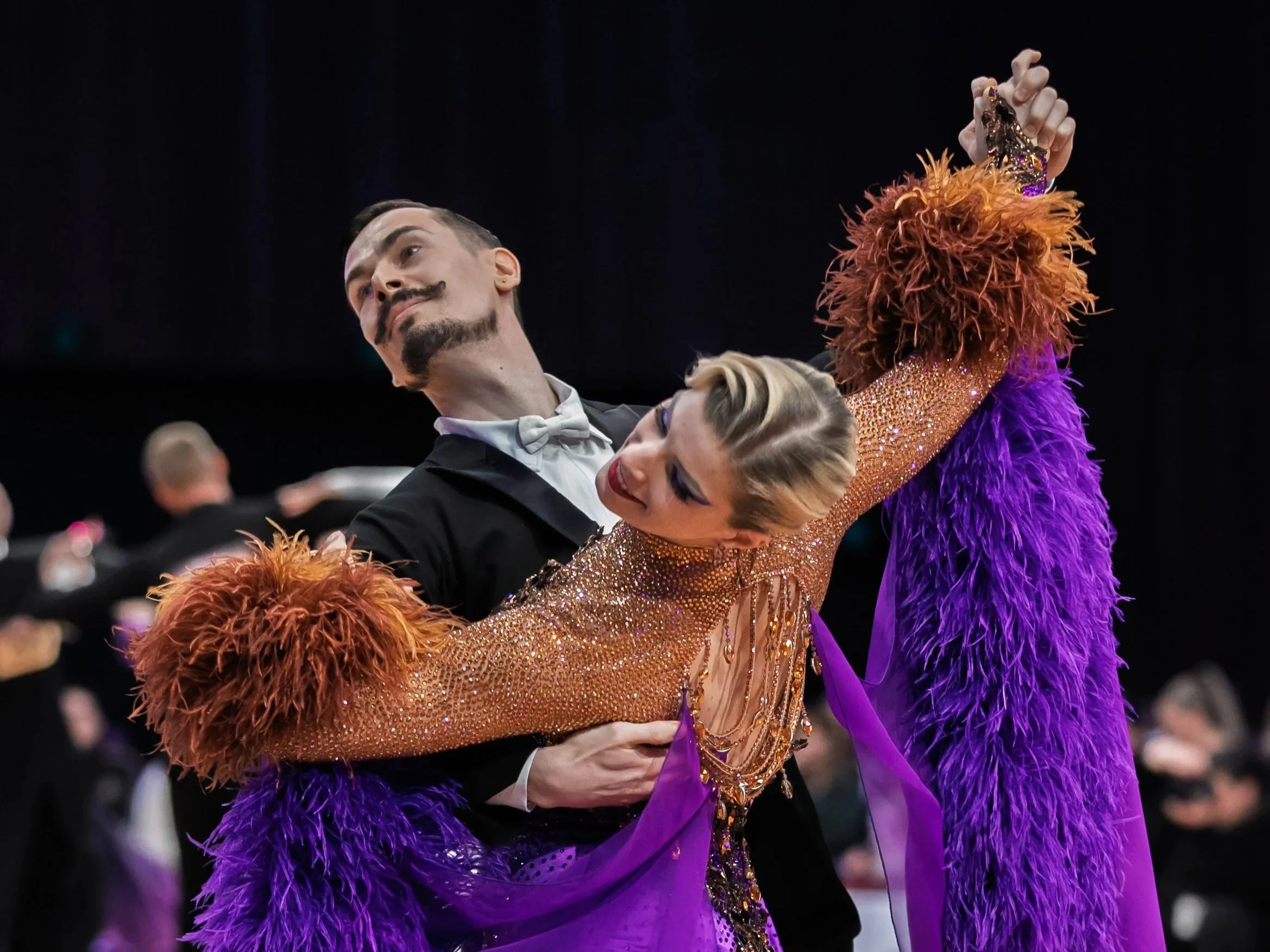 A couple dancing, with a man in a tuxedo and a woman in a glittery orange dress with purple feathers, at a formal event.