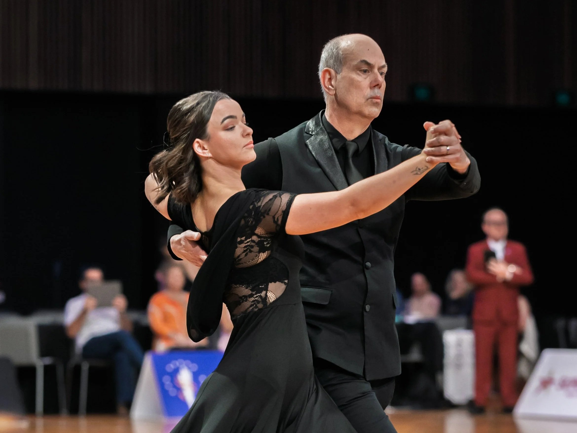 A man and woman dance closely, the man with his hand on the woman's back and their hands clasped together, both with serious expressions, in a ballroom with a dark background and blurred audience.