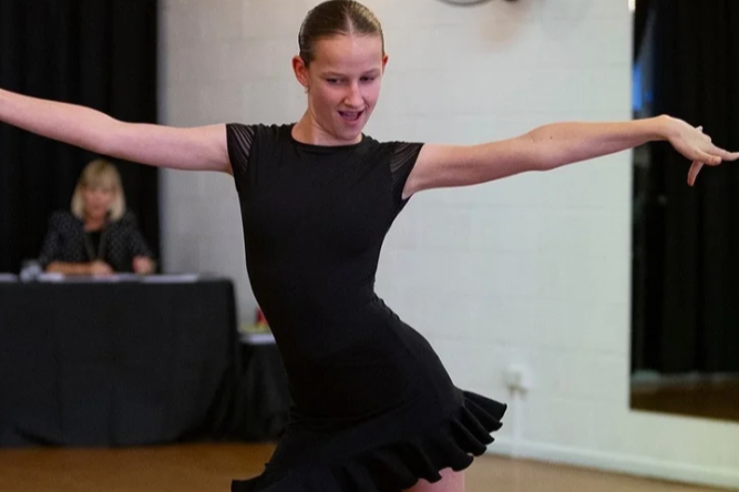 A young woman dancing, wearing a black dress, with her arms extended and a cheerful expression. In the background, a woman is seated at a table.