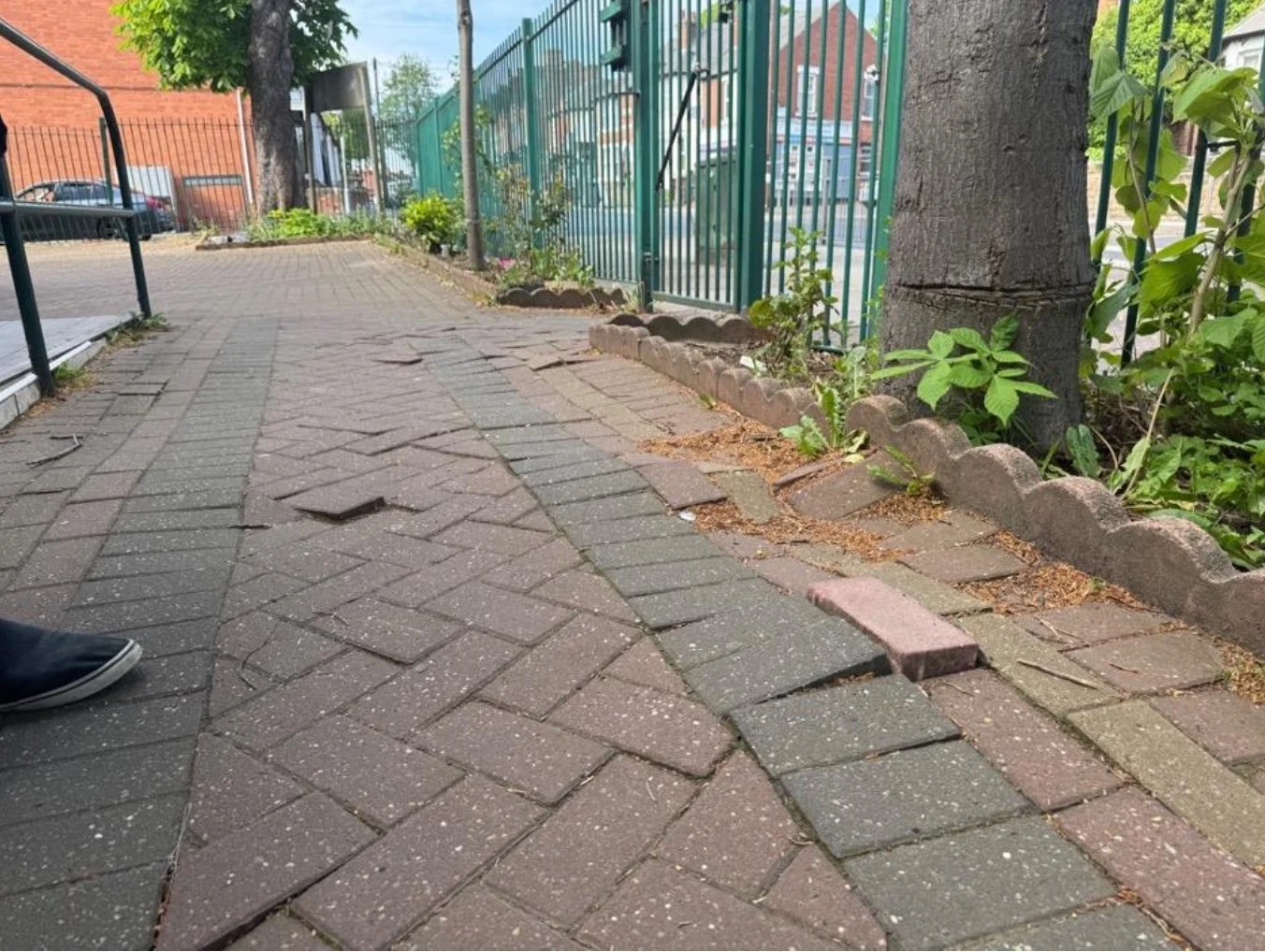 Broken brick pavement on a sidewalk with a green metal fence and trees in the background.