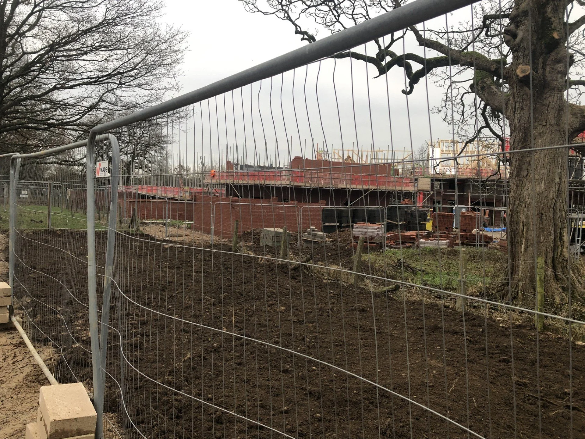 Construction site with fenced-off area, brick structures, and unfinished building in the background. Overcast sky with leafless trees nearby.