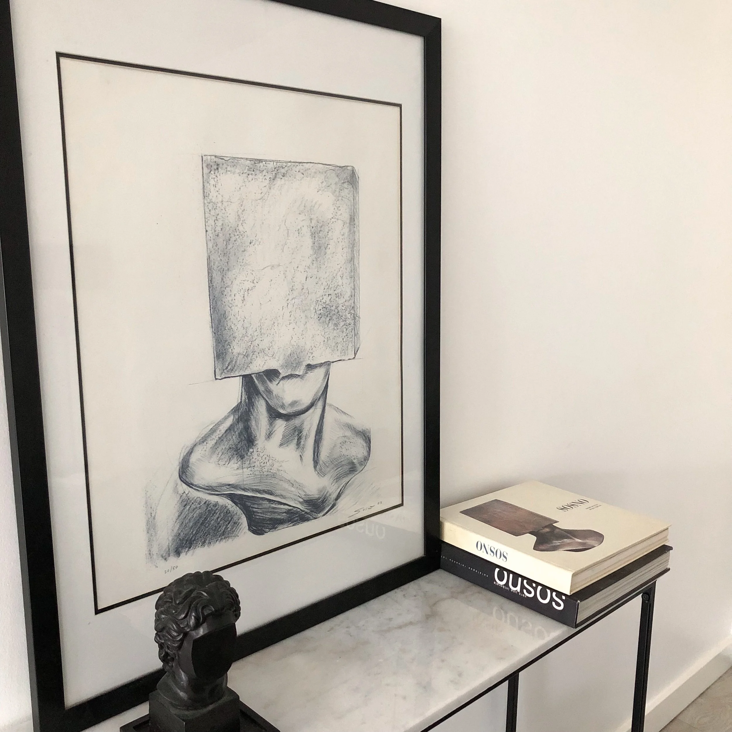 Black framed post-war lithographie of classical sculpture by sacha sosno. The sculpture is of a human head with a detailed curly hairstyle. The artwork is displayed on a white wall with a matte white surface showing at the bottom of the frame.