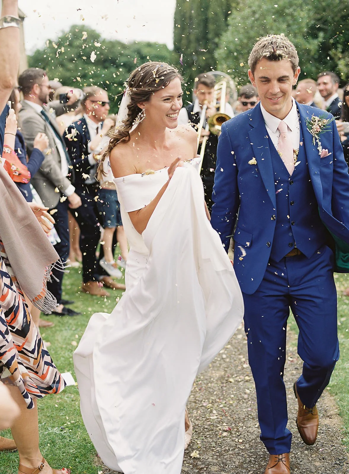 Bride and groom walking outdoors with wedding guests, confetti falling, green trees in background.