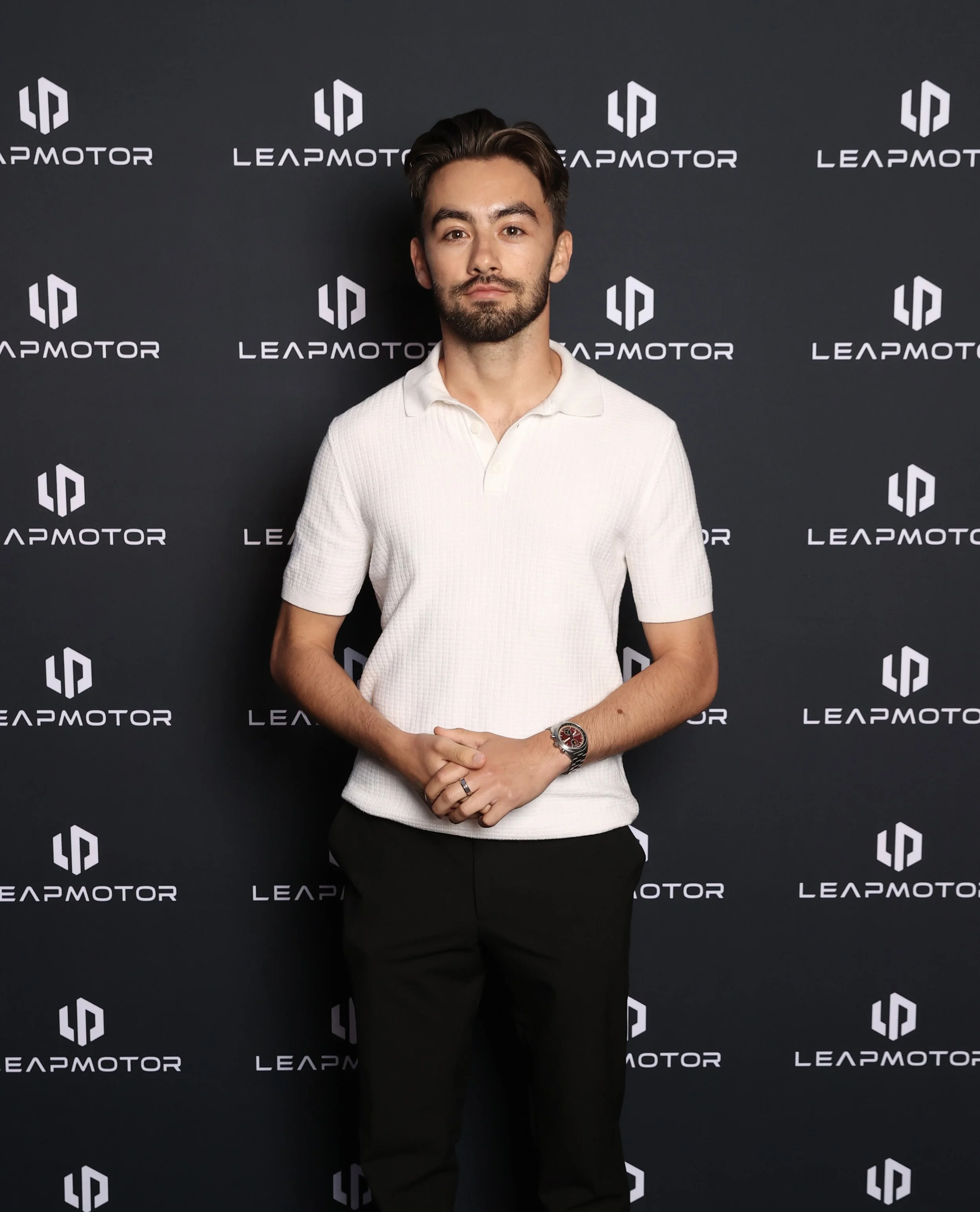 A man with dark hair, a beard, wearing a white polo shirt and black pants, standing with his hands clasped in front of him against a black backdrop with the words 'LEAPMOTOR' and its logo repeated.